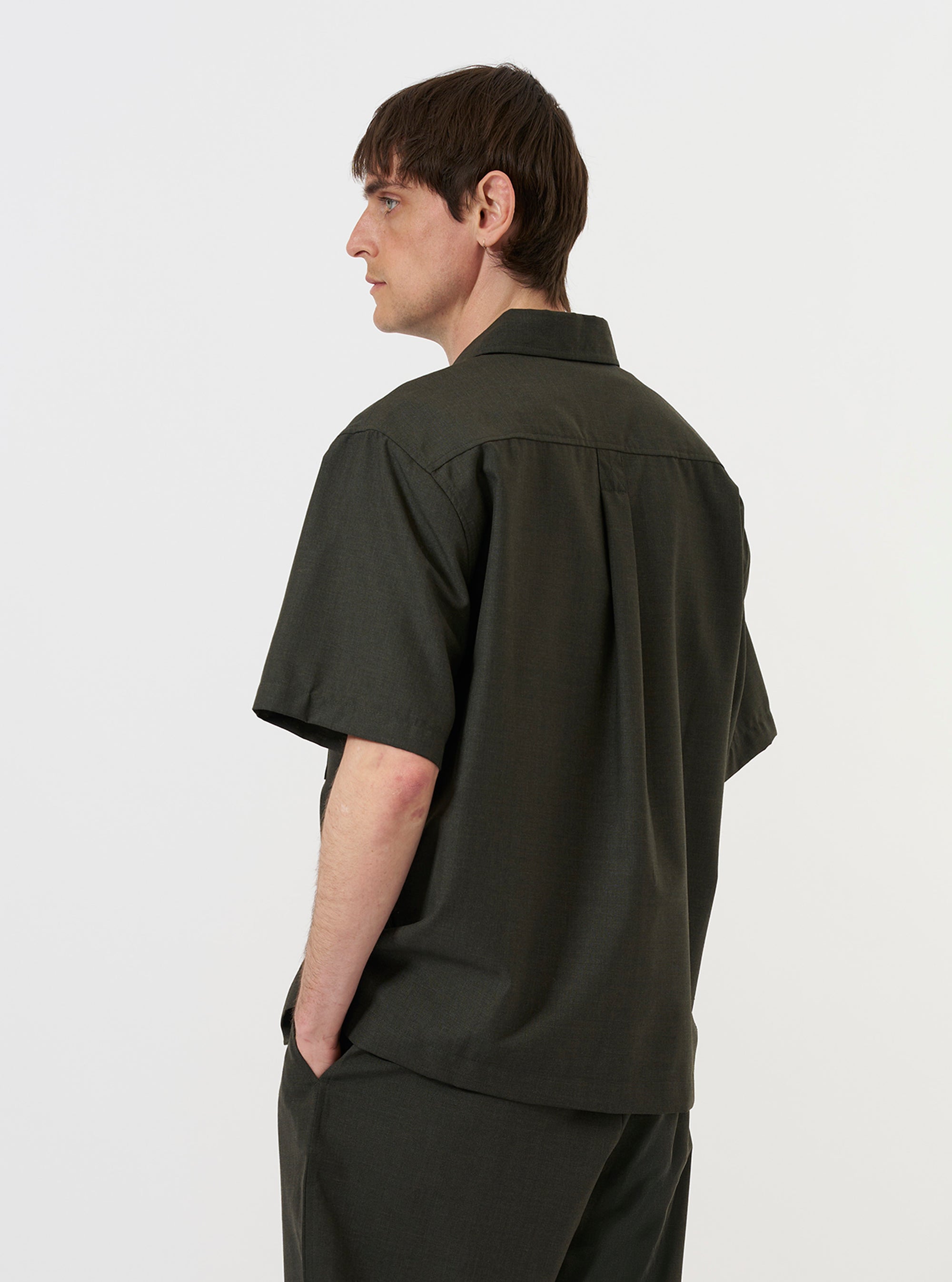 A model is wearing the Olive Marl Tropical Suiting Ewan Shirt against a plain white background. The image shows the garment from the back, highlighting the shirt's relaxed fit, the texture of the olive green marl fabric, and a simple pointed collar. The model is also wearing dark trousers, and the shirt is worn untucked. No mannequin is present in the image.