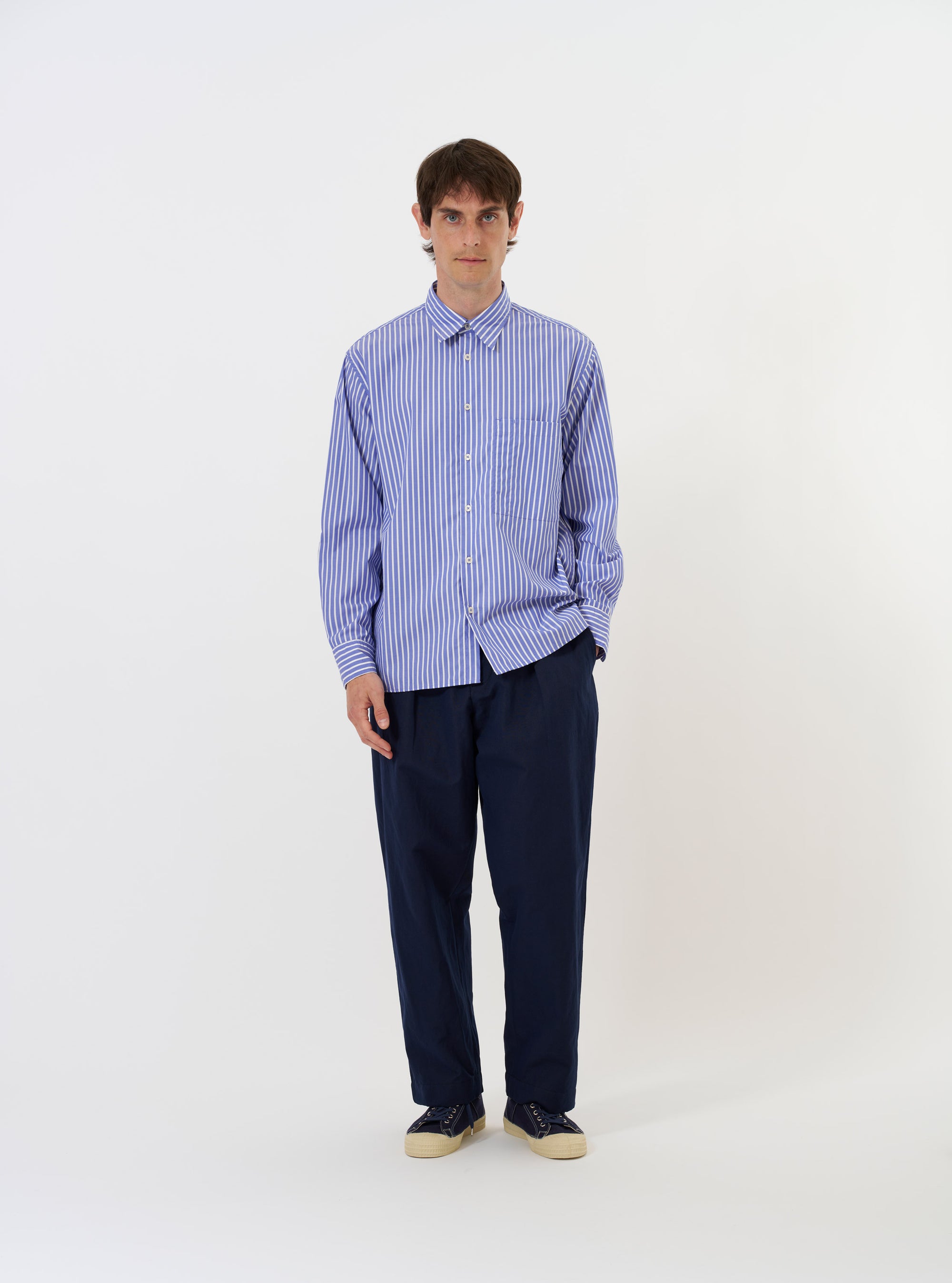 The Blue/White Cotton Stripe Square Pocket Shirt is shown laid flat against a solid white background. The garment features a light blue and white vertical stripe pattern, a classic pointed collar, and a full button-down front. A prominent, oversized square patch pocket is positioned on the left chest. The shirt has long sleeves with buttoned cuffs and a slightly curved hem.