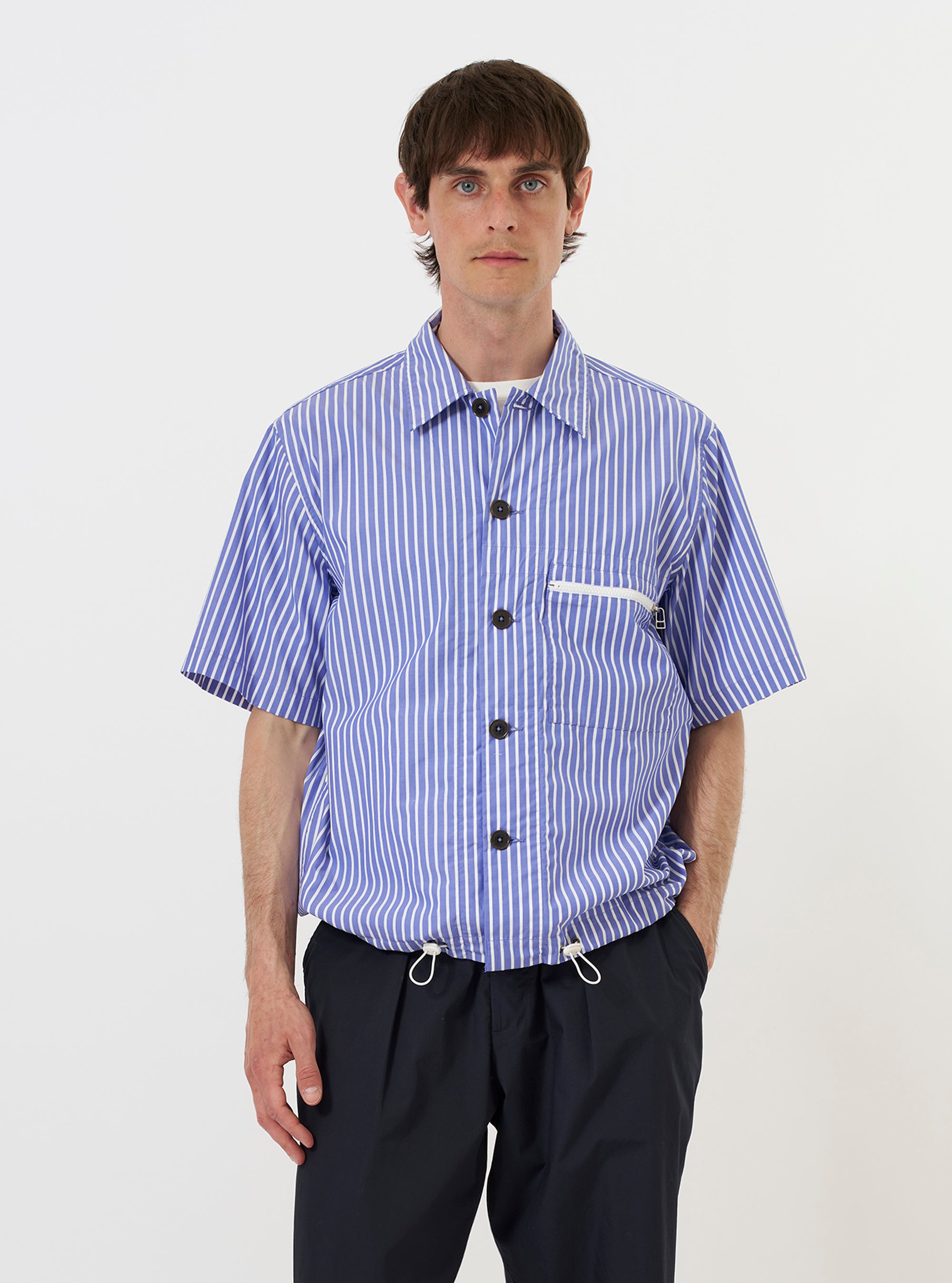 A model is wearing the Blue/White Cotton Stripe Z Tech Overshirt against a plain white background. The overshirt features a vertical blue and white stripe pattern, a single chest pocket with a visible zipper, and a classic pointed collar. The model is shown from the chest up, highlighting the technical details and fabric texture of the garment.