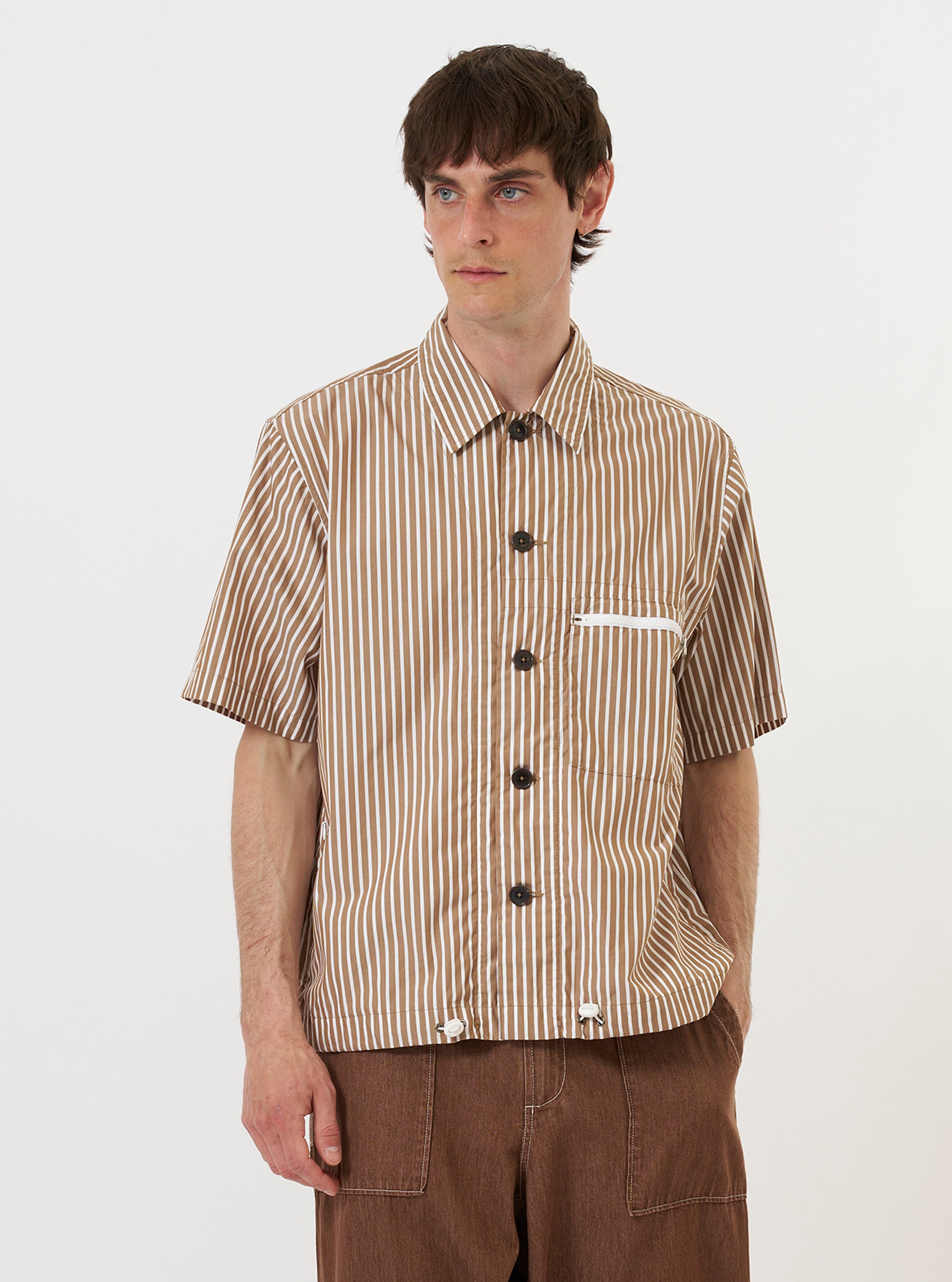 A model is wearing the Brown/White Cotton Stripe Z Tech Overshirt against a plain white background. The overshirt features a brown and white vertical stripe pattern, a classic pointed collar, and a full button-down front. A prominent zipped patch pocket is visible on the chest. The model is shown from the chest up, wearing the garment over a white t-shirt.