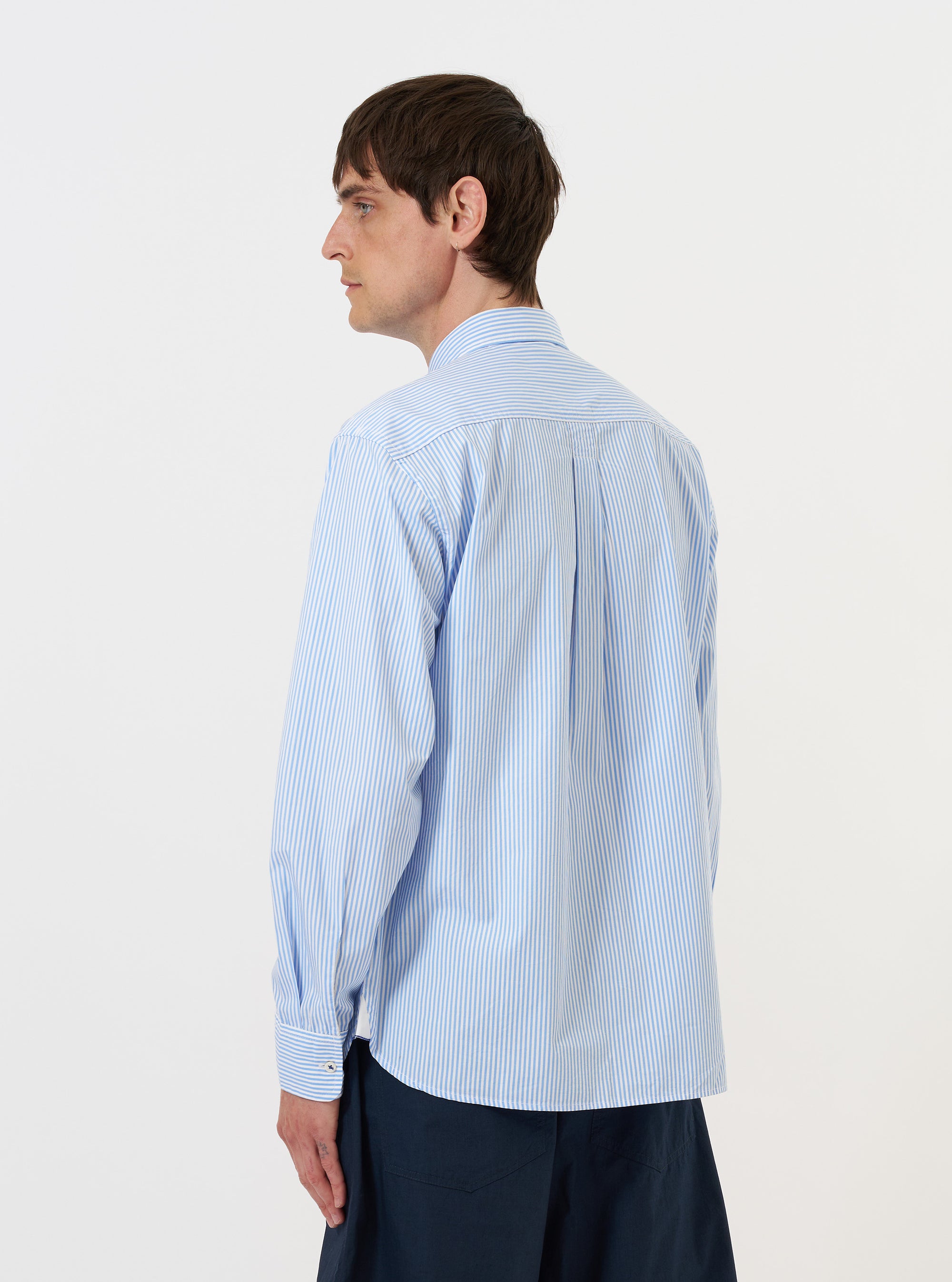 A model stands against a plain white background wearing the Sky/White Classic Nipon Stripe Lazy Day Shirt. The image shows the shirt from the back, highlighting the back yoke, a central box pleat, and the light blue and white vertical stripe pattern. The model is wearing the shirt untucked, paired with dark navy trousers, and has their hands in their pockets.