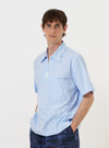 A model is wearing the Sky/White Classic Nipon Stripe Ewan Shirt against a plain white background. The shirt features vertical sky blue and white stripes, a quarter-zip neckline, and short sleeves. The model is shown from the chest up with the zipper partially open, highlighting the relaxed fit and the pattern of the garment.