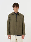 A model is wearing the Olive Linen Cotton Shirting Bakers Overshirt against a plain white background. The olive green garment is shown from the chest up, worn over a white t-shirt. The image highlights the one-piece collar and the textured linen-cotton fabric. The model is facing forward with a neutral expression.