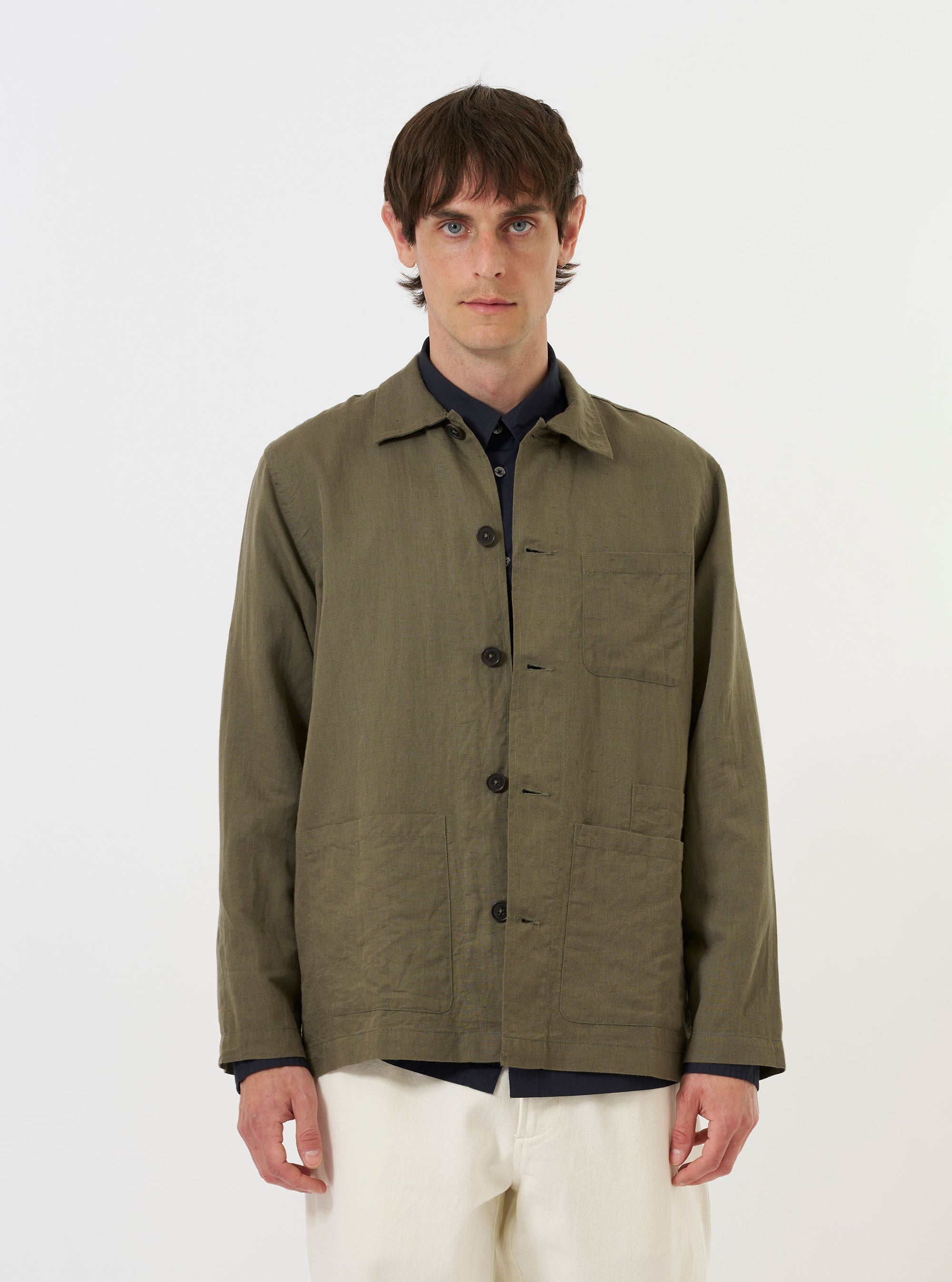 A model is wearing the Olive Linen Cotton Shirting Bakers Overshirt against a plain white background. The olive green garment is shown from the chest up, worn over a white t-shirt. The image highlights the one-piece collar and the textured linen-cotton fabric. The model is facing forward with a neutral expression.