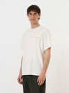 A model is wearing the Ecru Single Jersey Wish Wish You Were Here Tee against a plain white background. The image shows the front of the ecru-colored, short-sleeved t-shirt, which features a small graphic print on the left chest and a simple crew neck.
