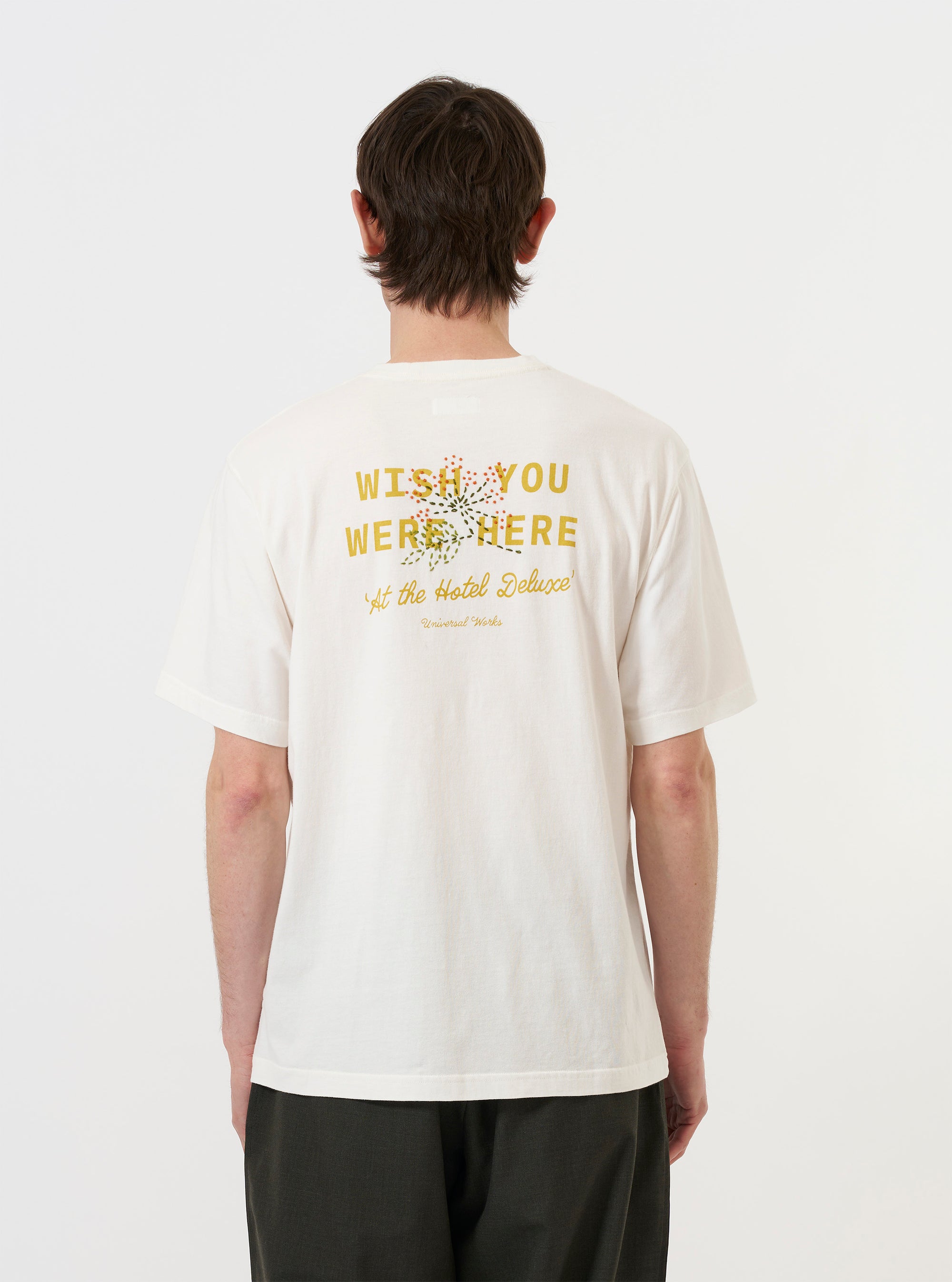 A model is seen from the back wearing the Ecru Single Jersey Wish Wish You Were Here Tee against a plain white background. The image highlights a large back graphic featuring the text "WISH YOU WERE HERE" above a floral spray print with hand-embroidered details. The ecru-colored t-shirt has a generous fit and short sleeves.