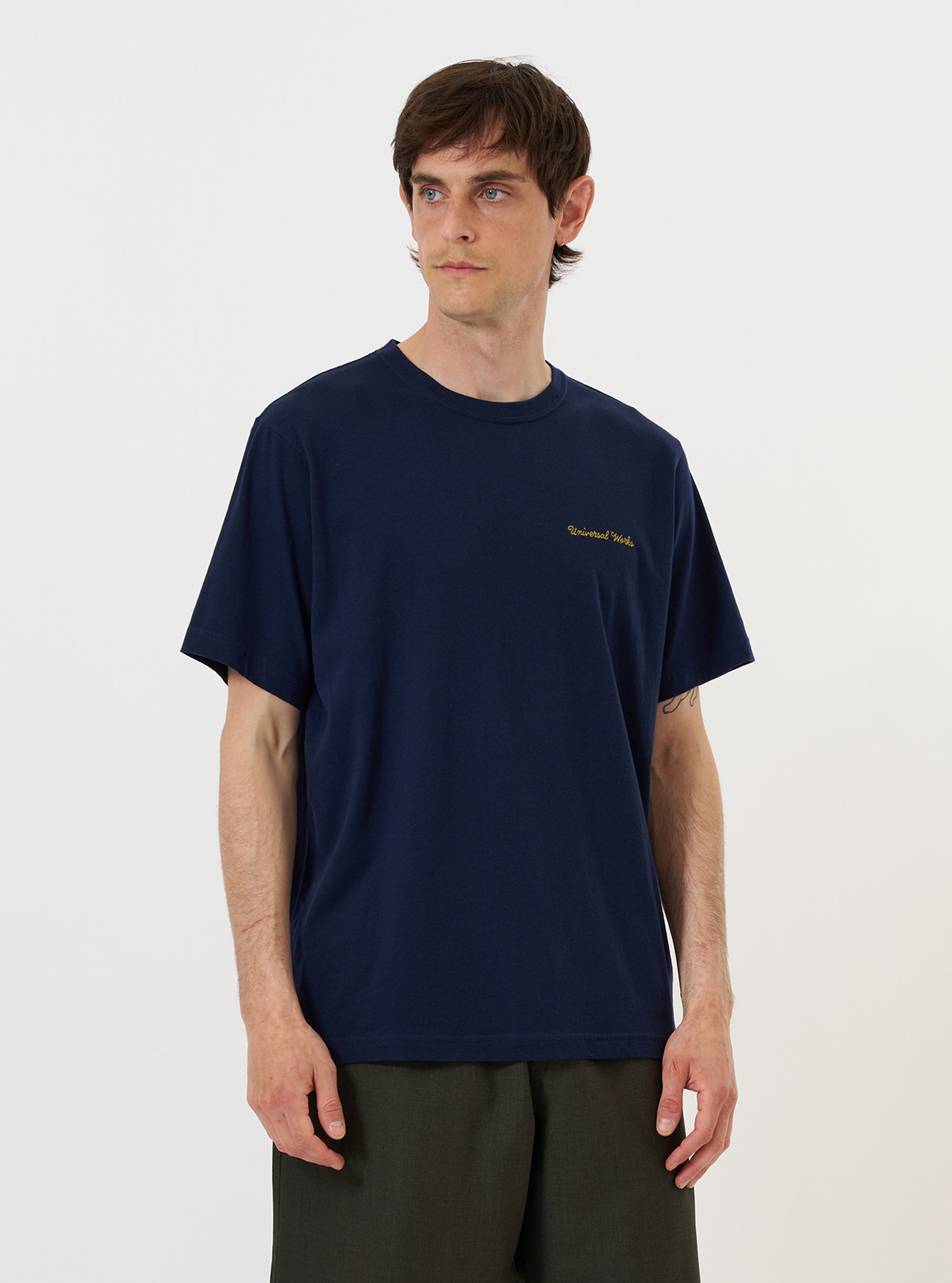A model is wearing the Navy Single Jersey Wish Wish You Were Here Tee against a plain white background. The navy blue t-shirt features a small white graphic on the left chest and is styled with olive green trousers. The image shows the model from the waist up, highlighting the regular fit and ribbed crew neck.