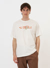 A model is wearing the Ecru Single Jersey Trail Trail To Hotel Delux Tee against a plain white background. The short-sleeved t-shirt features a relaxed fit and a large graphic print on the front with the words "Hotel Deluxe" and "Trail to Trail" alongside a mountain illustration. The model is standing and facing forward, showcasing the screen-printed design on the ecru-colored fabric.