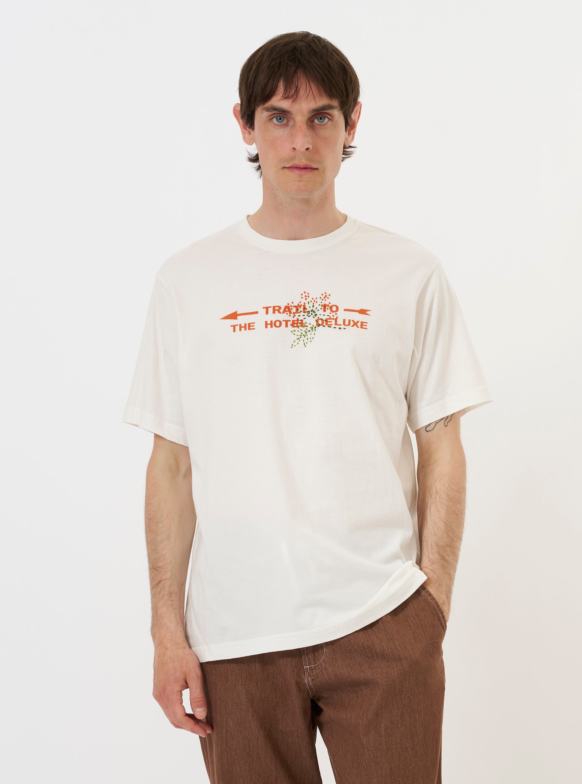 A model is wearing the Ecru Single Jersey Trail Trail To Hotel Delux Tee against a plain white background. The short-sleeved t-shirt features a relaxed fit and a large graphic print on the front with the words "Hotel Deluxe" and "Trail to Trail" alongside a mountain illustration. The model is standing and facing forward, showcasing the screen-printed design on the ecru-colored fabric.