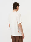 A model is seen from the back wearing the Ecru Single Jersey Trail Trail To Hotel Delux Tee against a plain white background. The ecru-colored t-shirt features a relaxed fit and a clean, straight hem. Only the upper half of the model is visible.