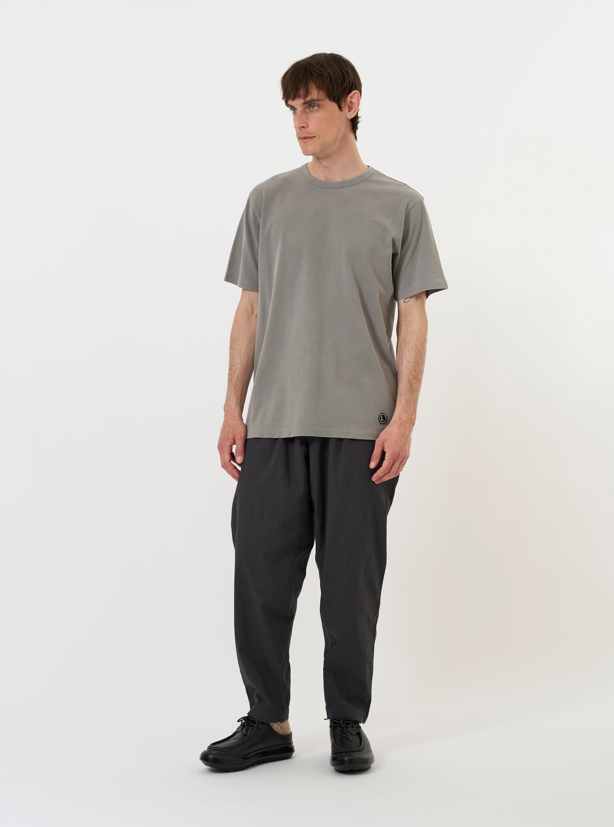 A mid grey piquet cotton T-shirt is shown laid flat against a solid white background. The garment features a regular fit, short sleeves, a classic crew neck with a self-bound collar, and a straight hem. A small, woven branded label is visible on the lower left side of the front.
