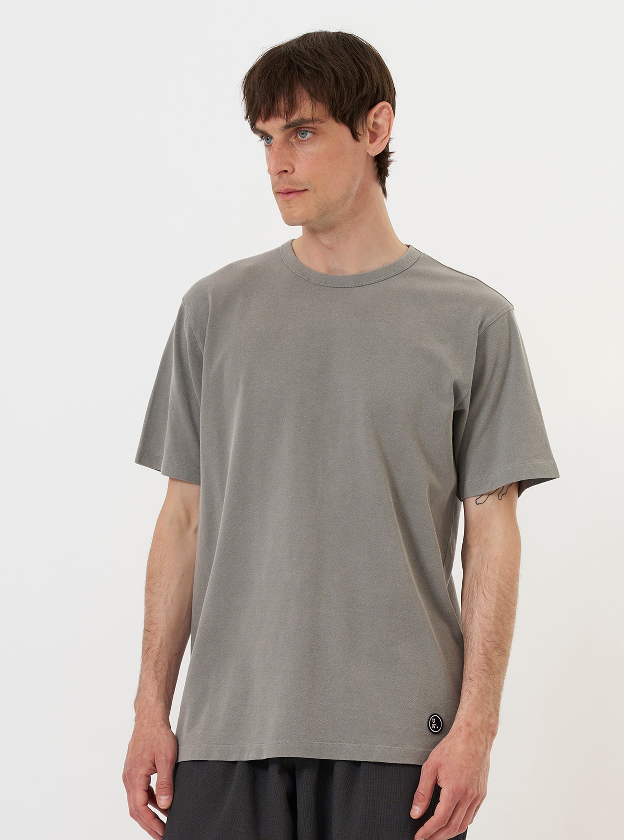 A model is wearing the Mid Grey Piquet Core Tee while standing against a plain white background. The mid-grey short-sleeved t-shirt features a subtle textured piquet weave and a ribbed crew neckline. The model is shown from the chest up, highlighting the fit and fabric texture of the garment.