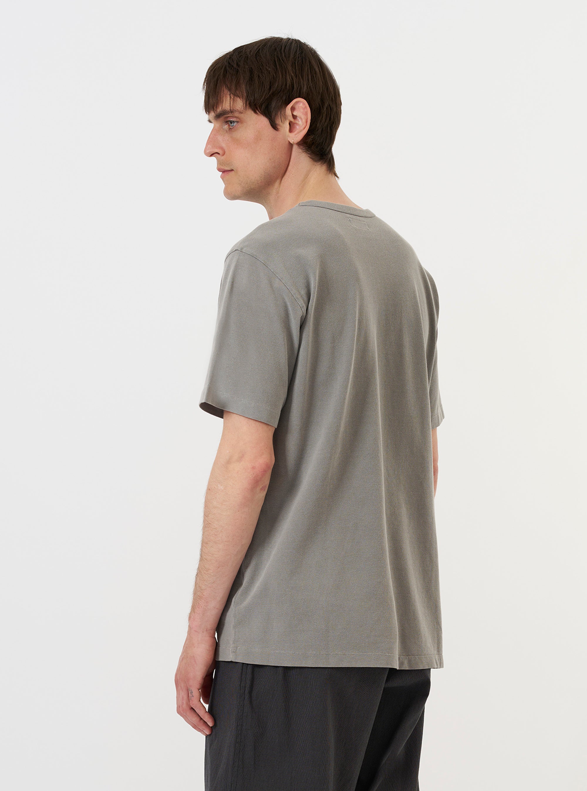 A model is wearing the Mid Grey Piquet Core Tee, standing against a plain white background. The image shows the back of the short-sleeved t-shirt, highlighting its clean, mid-grey piquet fabric and simple, straight hem. The model is wearing the garment untucked, paired with dark trousers.