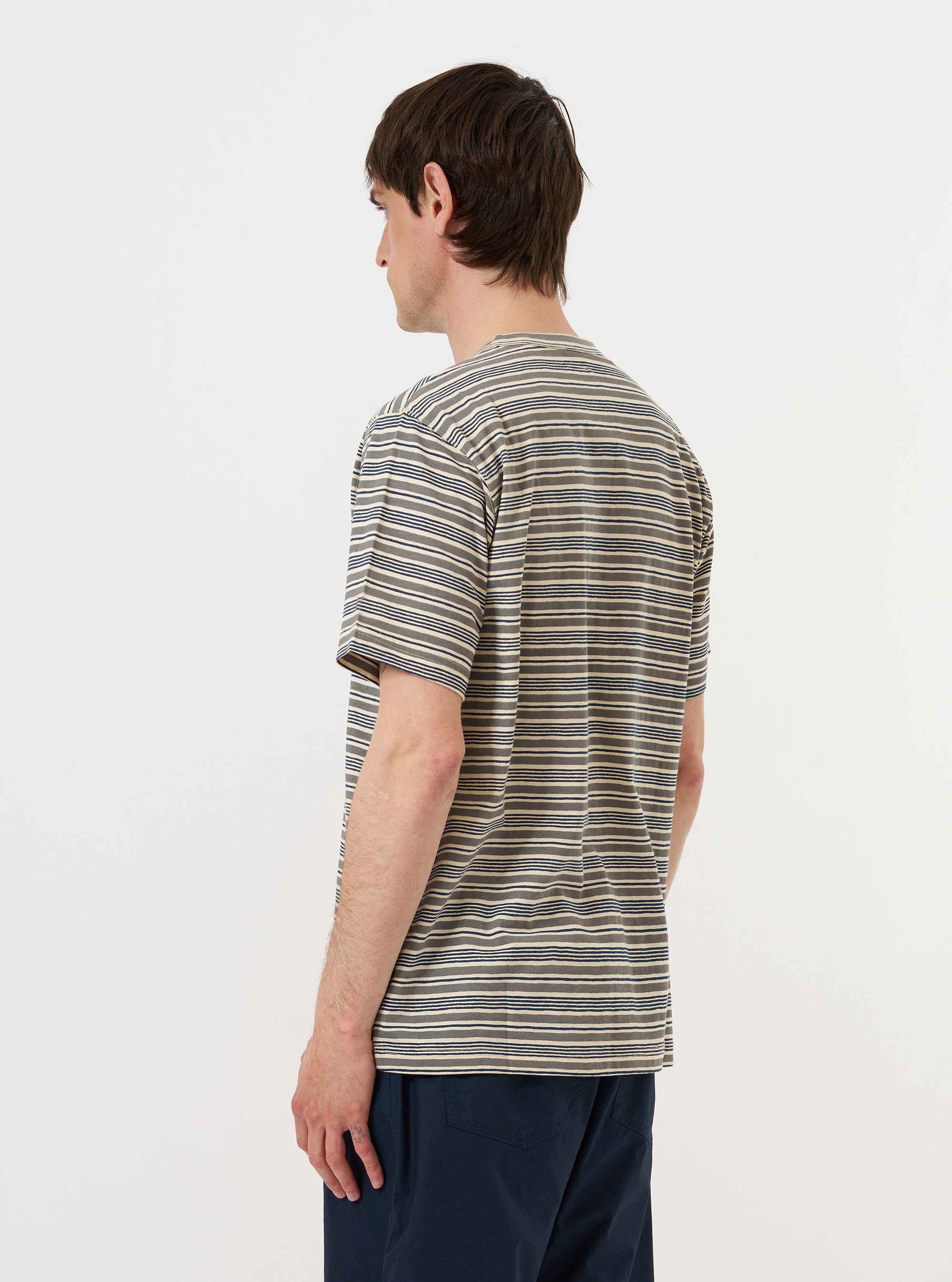 A model is seen from the back wearing the Grey OS Stripe Jersey Core Tee against a plain white background. The t-shirt features a horizontal grey and white stripe pattern, a relaxed oversized fit, and short sleeves. The image highlights the straight hem of the garment and the textured finish of the jersey fabric.