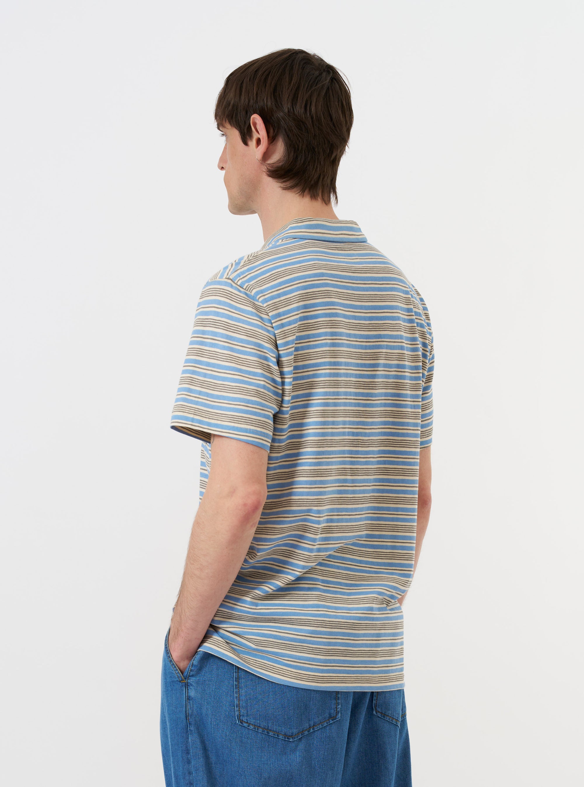 The Pale Blue OS Stripe Jersey Vacation Polo is displayed from a rear-side angle, laid flat against a plain white background. The image highlights the horizontal light blue and white stripes, the short sleeves, and the stepped hem with small side vents.