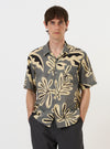 A model is wearing the Grey Baja Print Road Shirt against a plain white background. The short-sleeved shirt features a detailed all-over botanical print in shades of grey and white, a relaxed camp collar, and a single chest patch pocket. The garment is shown from the waist up, worn buttoned and untucked to highlight its regular fit and straight hem.