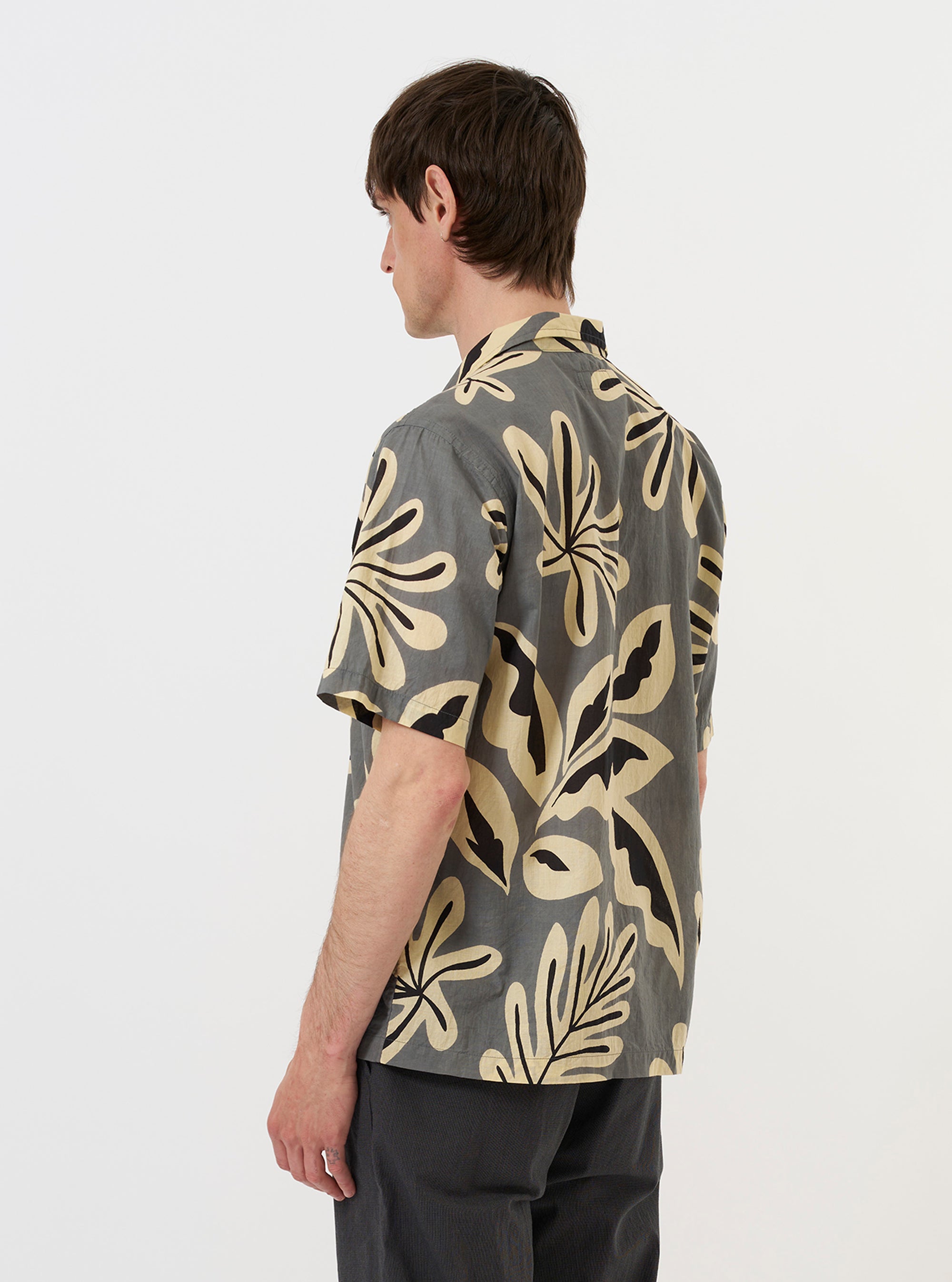 A model is wearing the Grey Baja Print Road Shirt against a plain white background. The shirt features a dark grey fabric with a white geometric Baja print, a relaxed camp collar, and a button-down front. The model is shown from a side-rear perspective, highlighting the garment's fit and the continuous print pattern across the back and shoulders.