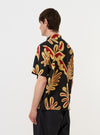 A model is seen from the back wearing the Black Baja Print Road Shirt against a plain white background. The shirt features a black base with a white geometric Baja print, short sleeves, and a straight hem. The model has their hands in their pockets, highlighting the relaxed fit of the garment.