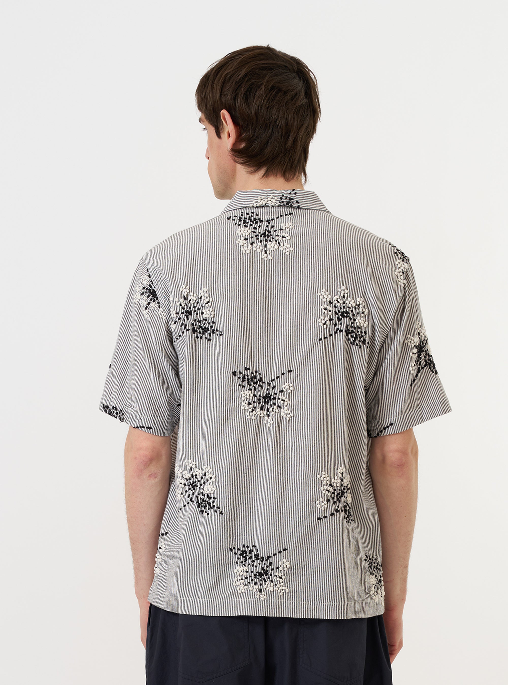 A model is wearing the Grey Hand Embroidered Stripe Cotton Road Shirt against a plain white background. The image provides a rear view of the shirt, showcasing the hand-embroidered vertical stripes on the grey cotton fabric, the short sleeves, and the straight hem. The model is standing and pairing the shirt with dark trousers.