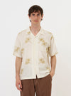 A male model is wearing the Ecru Hand Embroidered Cotton Road Shirt against a plain white background. The short-sleeved shirt features a relaxed fit, a camp collar, and a single chest patch pocket, with dark floral embroidery motifs visible on the front and sleeves. The model is standing and facing forward, with the shirt fully buttoned.