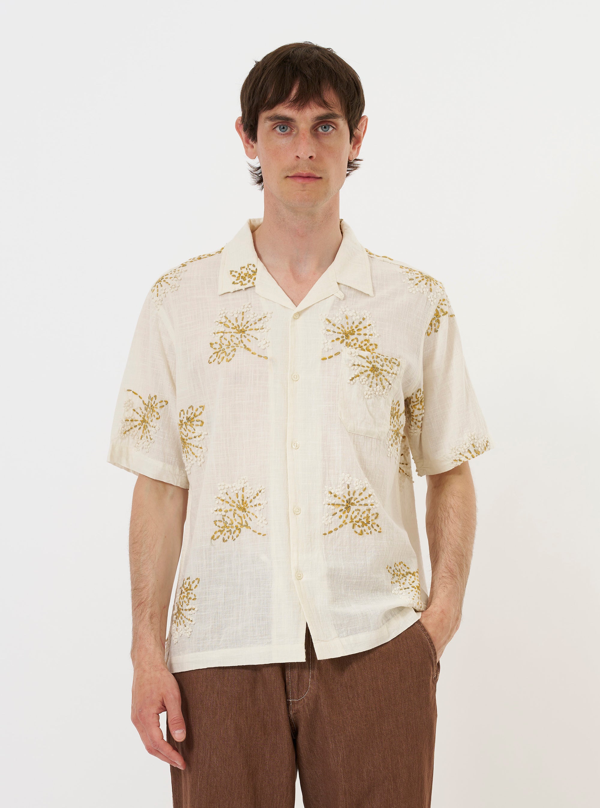 A male model is wearing the Ecru Hand Embroidered Cotton Road Shirt against a plain white background. The short-sleeved shirt features a relaxed fit, a camp collar, and a single chest patch pocket, with dark floral embroidery motifs visible on the front and sleeves. The model is standing and facing forward, with the shirt fully buttoned.