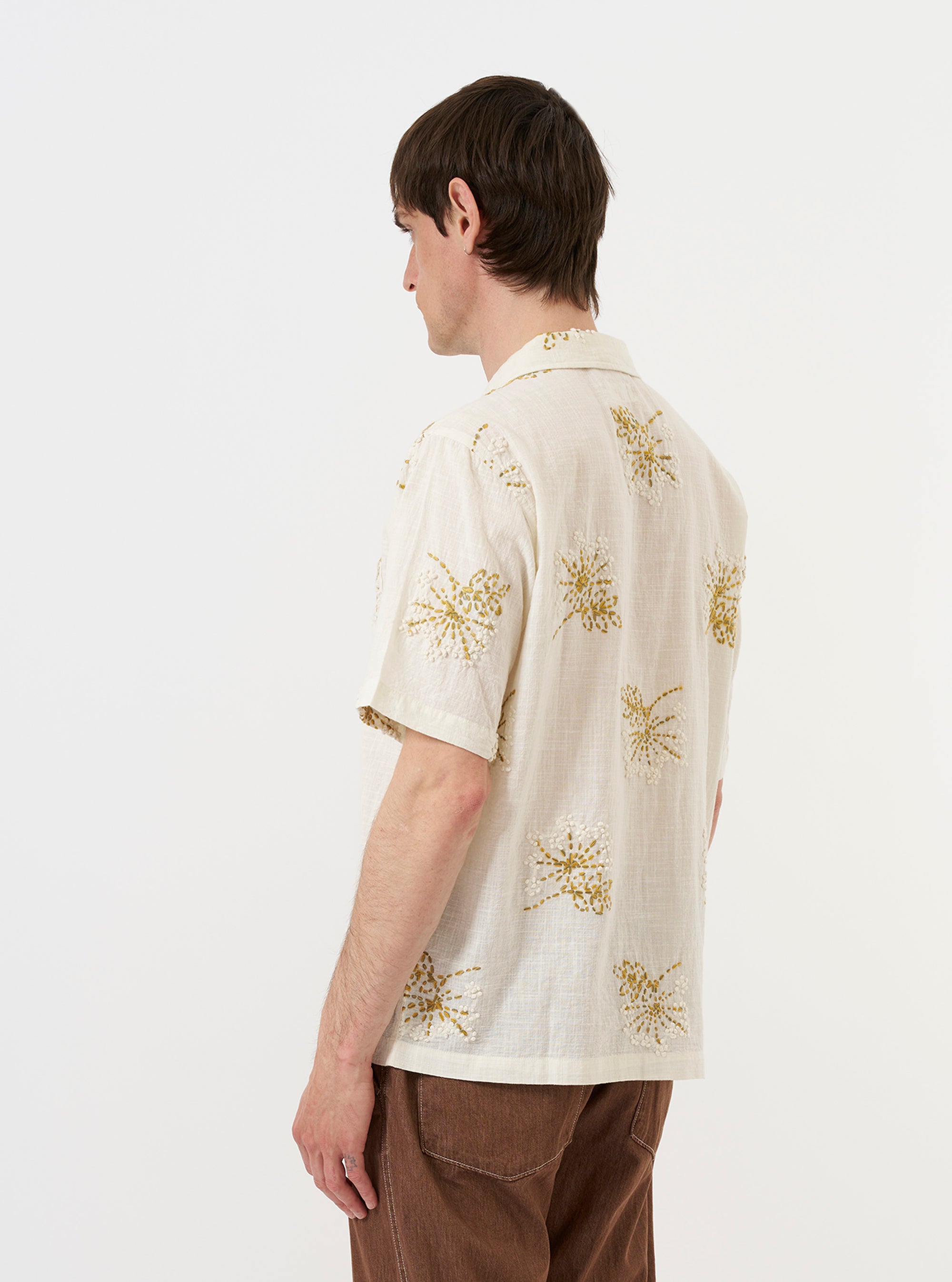 A male model is wearing the Ecru Hand Embroidered Cotton Road Shirt against a plain white background. The short-sleeved shirt features a camp collar, a single chest patch pocket, and a straight hem. The ecru fabric is detailed with subtle vertical stripes and small, colorful floral hand-embroidered motifs scattered across the garment. The model is shown from the torso up, facing forward.