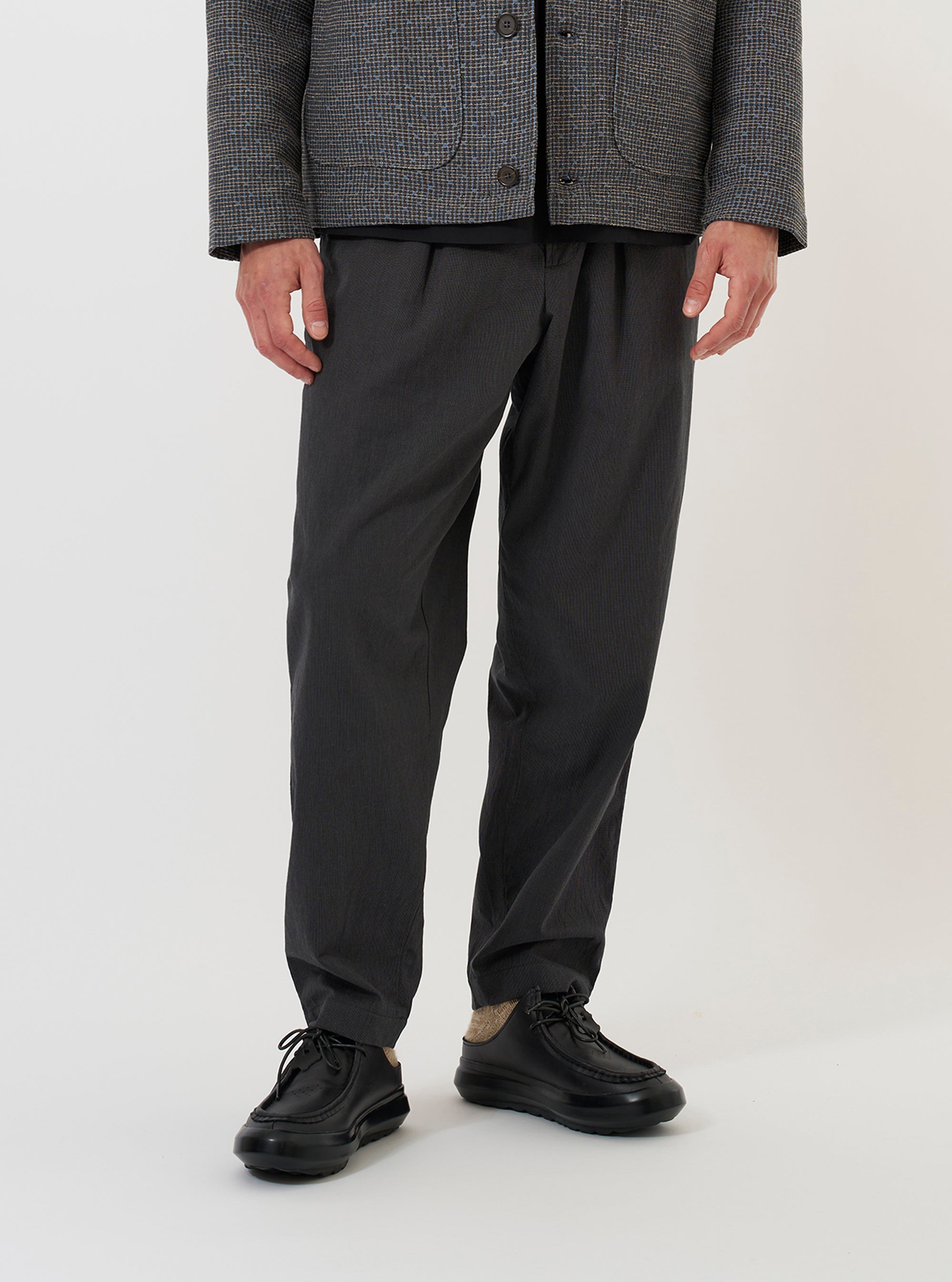 A model is wearing the Charcoal Seattle Cotton Pleated Track Pant against a plain white background. The charcoal grey trousers are shown from the waist down, featuring a loose, tapered fit with a single pleat at the waist and a drawstring waistband. The model is standing and has their hands in the side slip pockets.