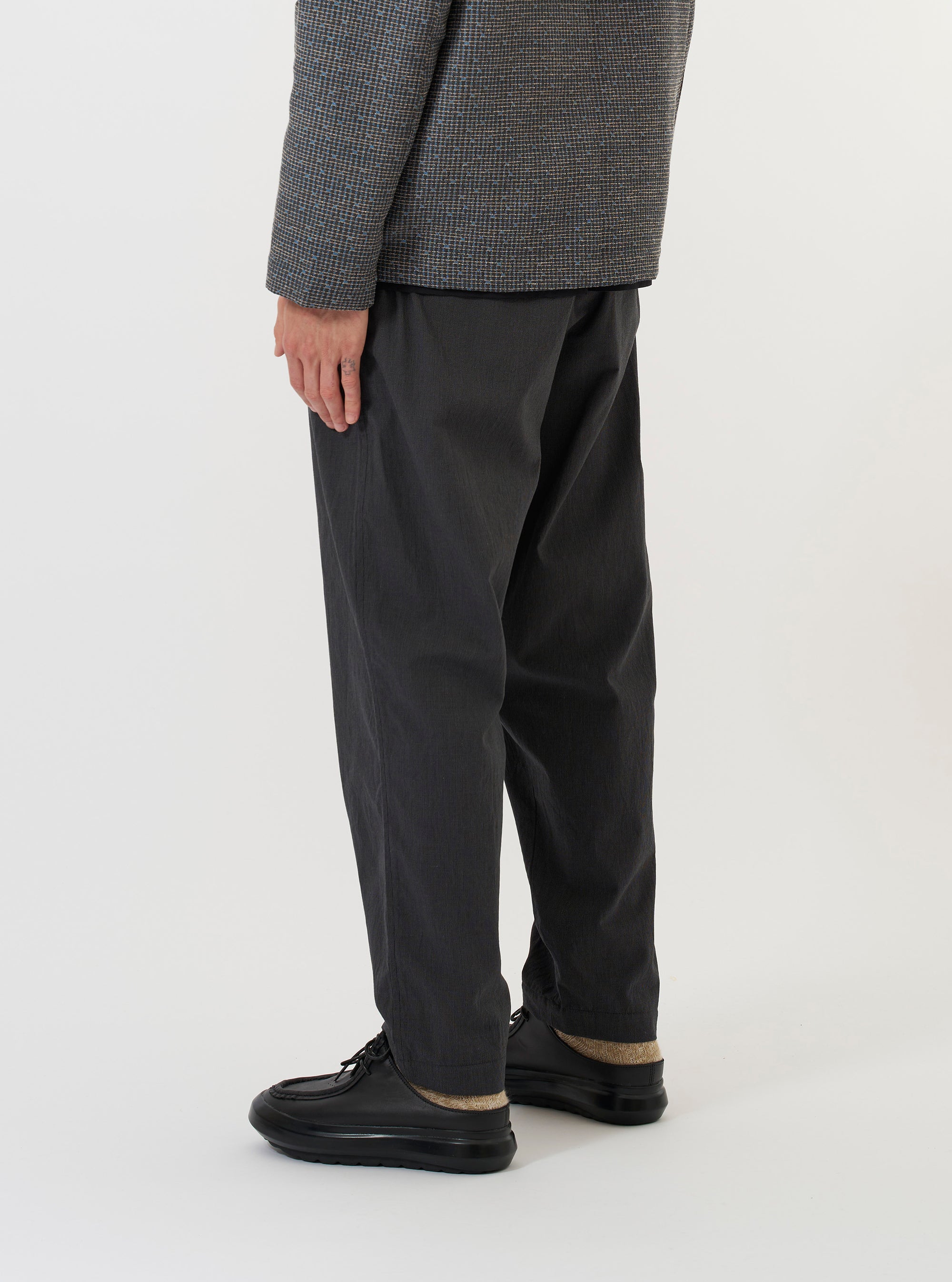 A model is wearing the Charcoal Seattle Cotton Pleated Track Pant against a plain white background. The charcoal grey trousers are shown from the waist down, highlighting the loose fit, tapered leg, and single pleat detail at the front. The elasticated waistband with a visible drawstring is also prominent as the model stands facing slightly to the side.