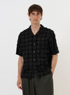 A model is wearing the Black 3D Check Road Shirt against a plain white background. The shirt features a textured black and grey 3D check pattern, a relaxed camp collar, and a button-down front. The model is shown from the waist up, wearing the shirt open over a dark undershirt, highlighting the garment's fit and the intricate weave of the fabric.