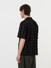 A model is wearing the Black 3D Check Road Shirt against a plain white background. The shirt features a black and grey textured 3D check pattern, a relaxed camp collar, and short sleeves. The model is shown from the back, highlighting the straight hem and the drape of the cotton fabric over the shoulders and torso. No mannequin is present in the image.
