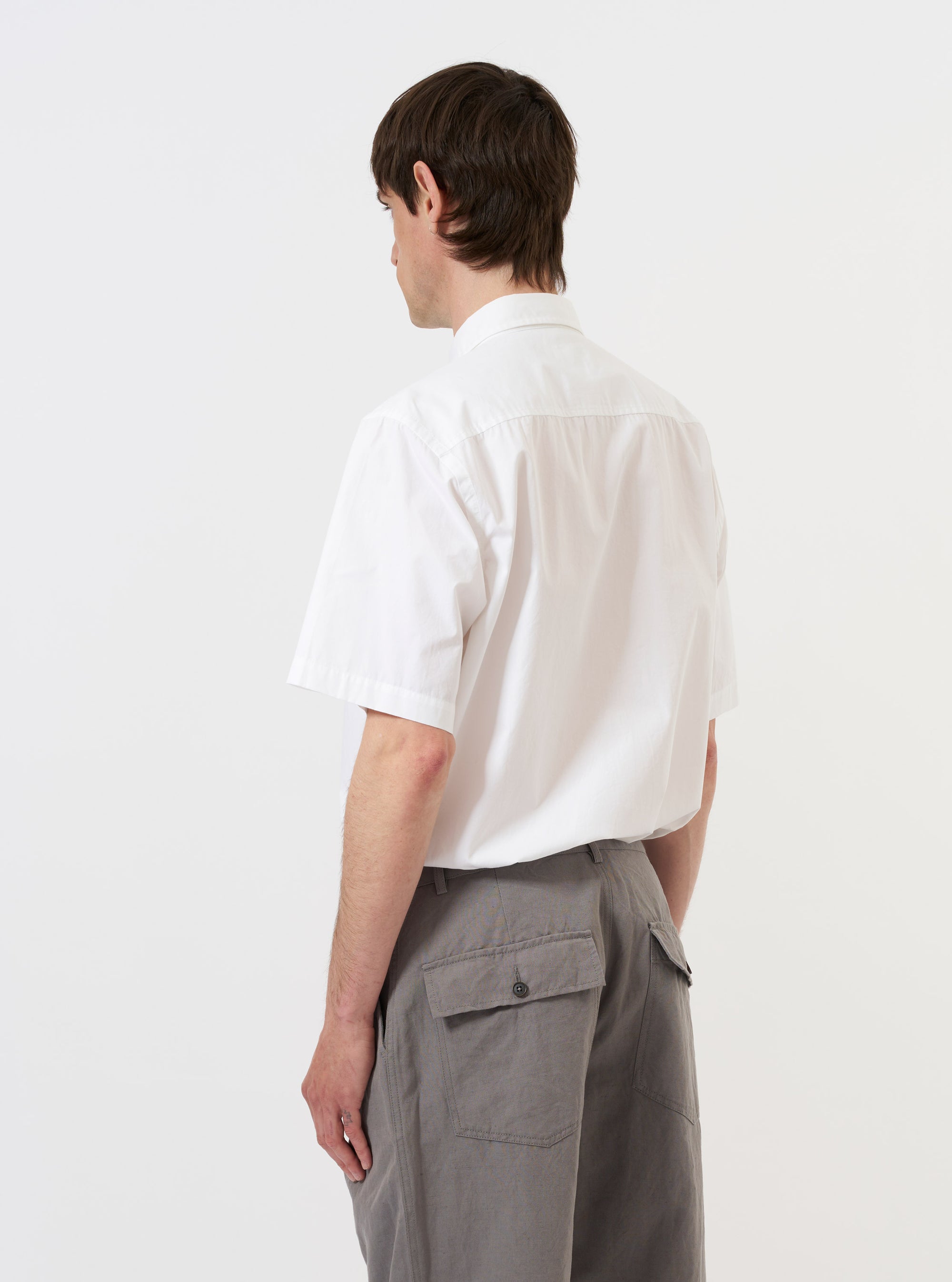 A model is seen from the back wearing the White Poplin Frill Shirt against a plain white background. The shirt features a clean white cotton poplin fabric with a prominent ruffled or frilled trim along the back yoke and shoulders. The model is also wearing dark trousers, and the image highlights the crisp texture and unique decorative detailing of the garment's rear construction. No mannequin is present in the image.