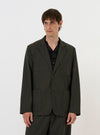A model stands against a plain white background wearing the Olive Marl Tropical Suiting Two Button Jacket. The jacket is an unstructured, relaxed fit in olive green marl, featuring a two-button front closure, a long, wide lapel, two lower patch pockets, and a single chest patch pocket. The model is shown from the waist up, layering the jacket over a simple top.