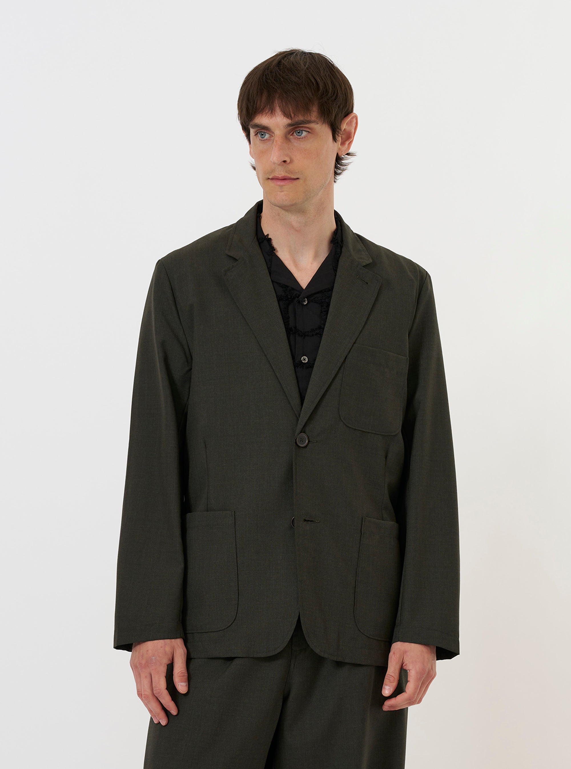 A model stands against a plain white background wearing the Olive Marl Tropical Suiting Two Button Jacket. The jacket is an unstructured, relaxed fit in olive green marl, featuring a two-button front closure, a long, wide lapel, two lower patch pockets, and a single chest patch pocket. The model is shown from the waist up, layering the jacket over a simple top.