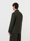 A model is wearing the Olive Marl Tropical Suiting Two Button Jacket against a plain white background. The image, taken from a side-rear perspective, highlights the jacket's structured shoulder, the back vent, and the olive green marl texture of the fabric.