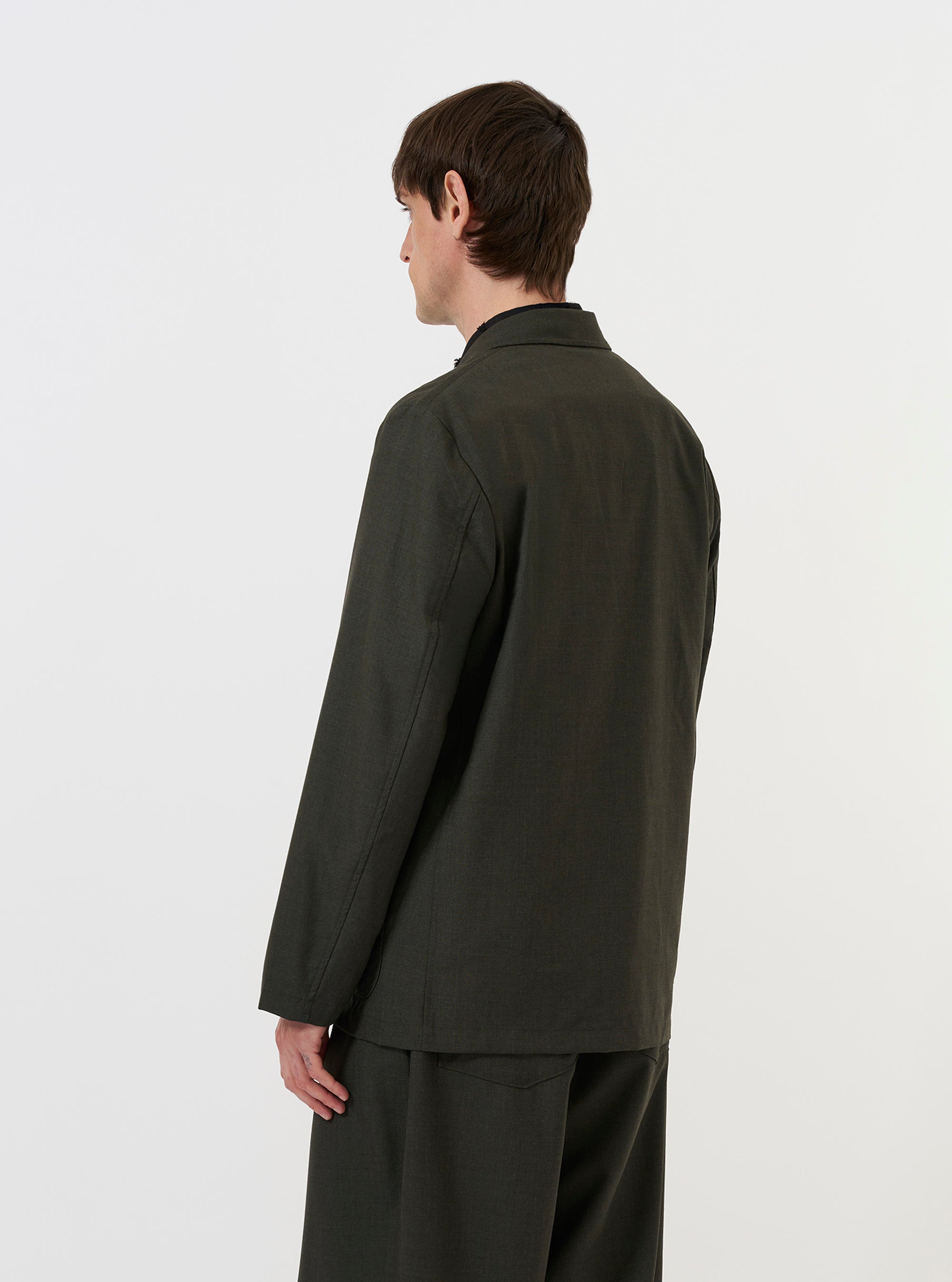 A model is wearing the Olive Marl Tropical Suiting Two Button Jacket against a plain white background. The image, taken from a side-rear perspective, highlights the jacket's structured shoulder, the back vent, and the olive green marl texture of the fabric.