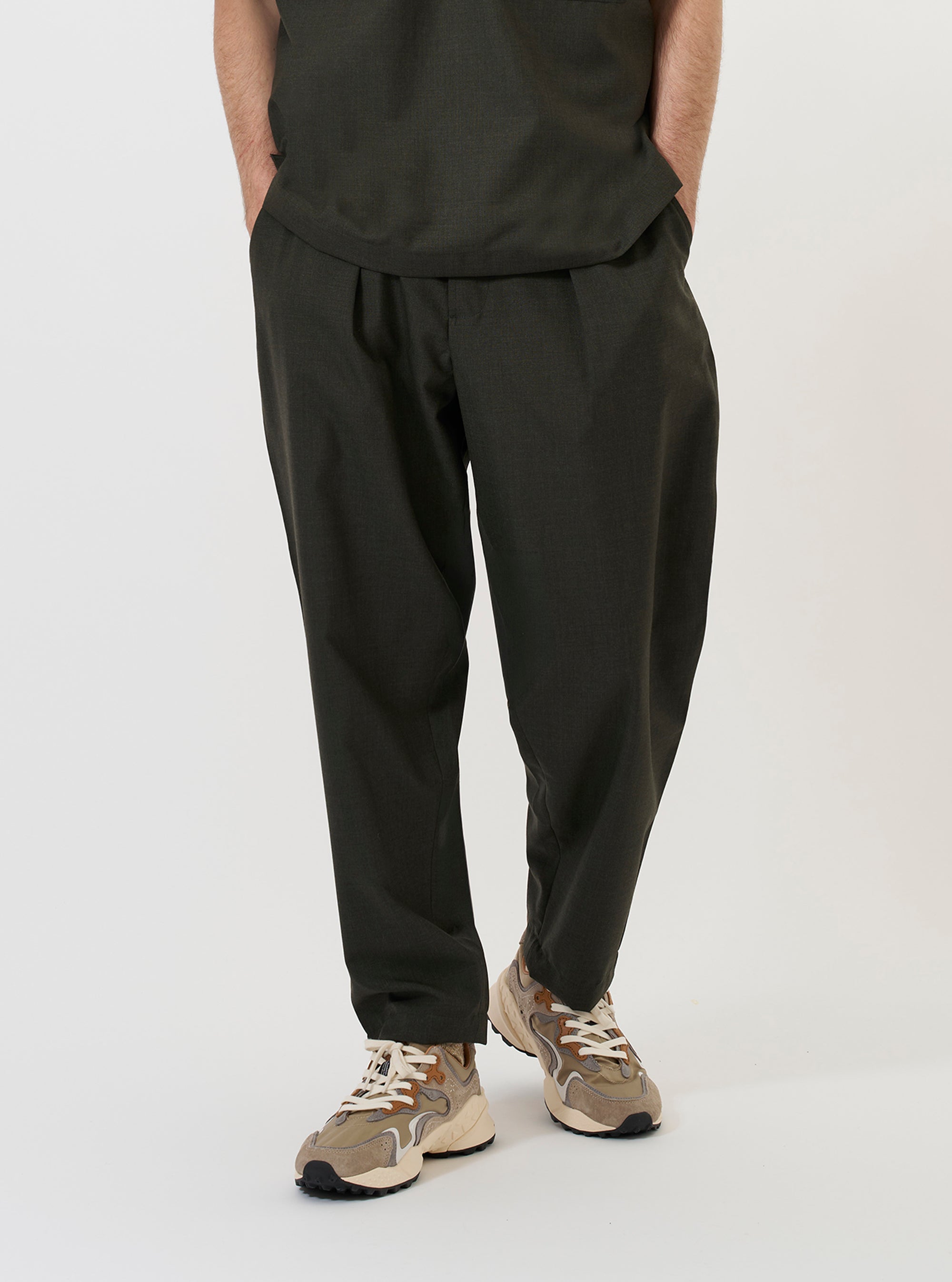A model is wearing the Olive Marl Tropical Suiting Pleated Track Pant against a plain white background. The image shows the model from the waist down, highlighting the loose, tapered fit of the olive green trousers. Key details visible include the single pleat at the front, side slip pockets, and the drape of the tropical suiting fabric. The model is standing and is also wearing dark shoes.