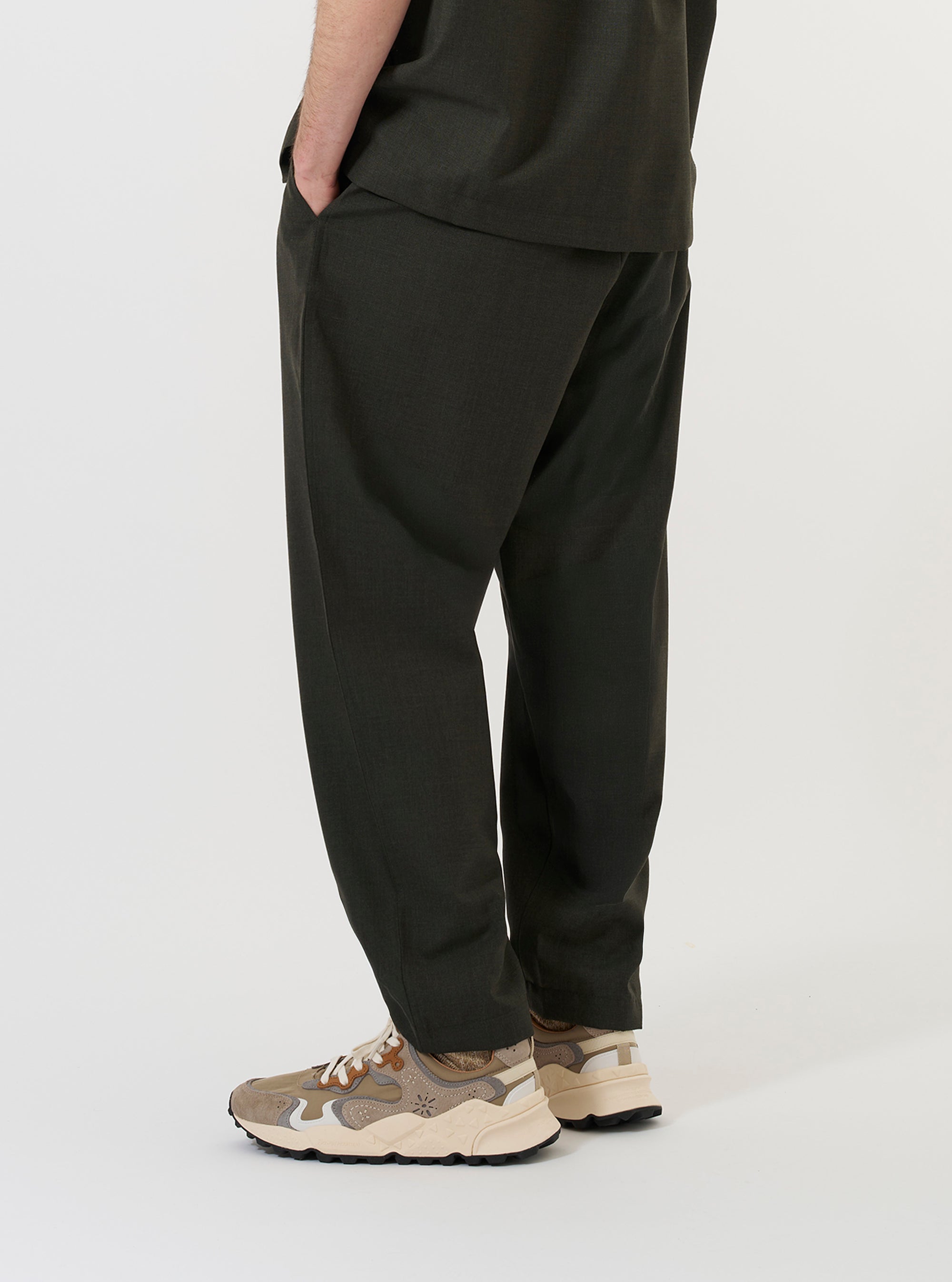 A model is shown from the waist down wearing the Olive Marl Tropical Suiting Pleated Track Pant against a plain white background. The image provides a side-rear perspective of the olive green trousers, highlighting the loose, tapered fit and the texture of the tropical suiting fabric. The model is standing, showcasing the relaxed drape of the garment.