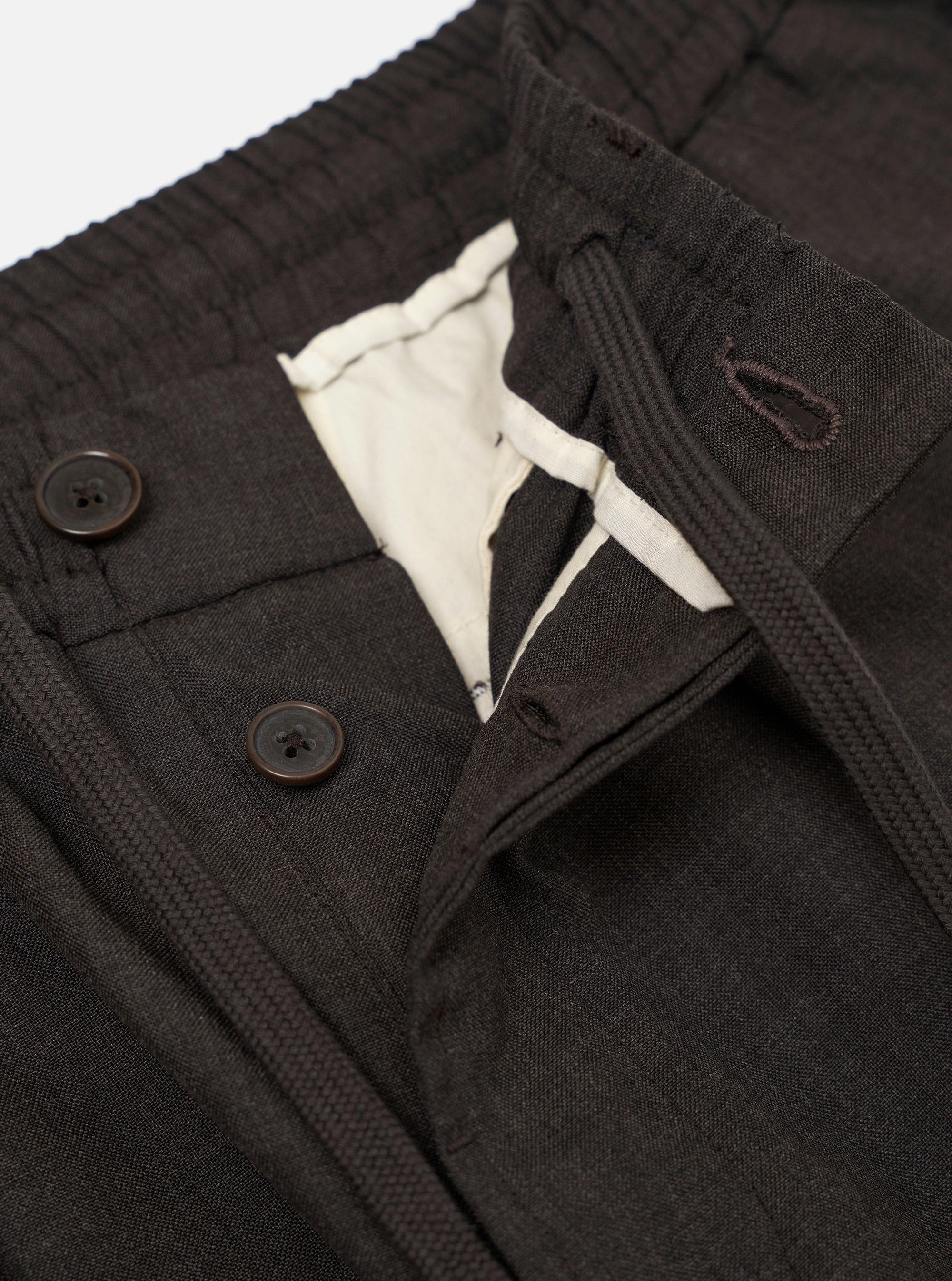A pair of brown marl tropical suiting pleated track pants is displayed flat against a plain white background. The image highlights the loose, tapered fit and the textured, flecked appearance of the wool-blend fabric. Key details include an elasticated waistband with an adjustable drawstring and a single pleat at the front.