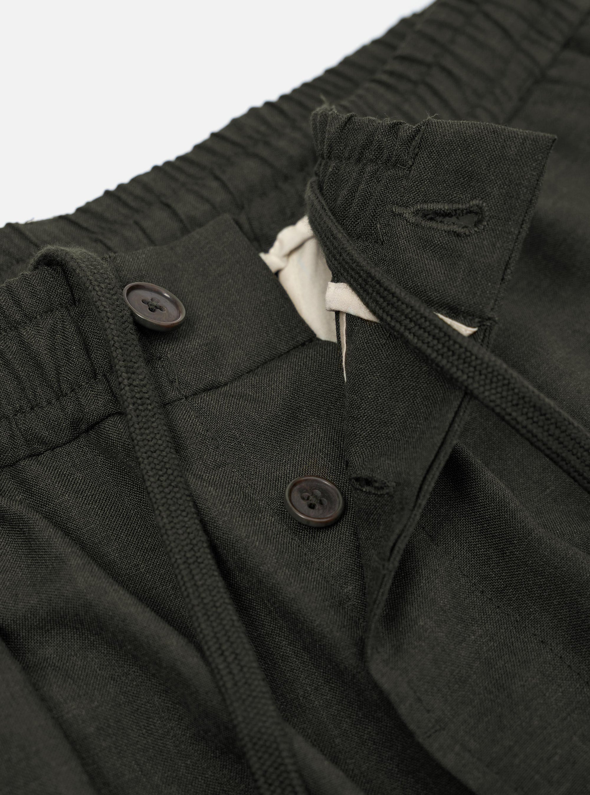 A pair of Olive Marl Tropical Suiting Pleated Track Pants is shown from the back, laid flat against a plain white background. The trousers feature a loose, tapered leg and an elasticated waistband. Key details include two distinct rear pockets, one with a buttoned flap and one jetted with a button closure.