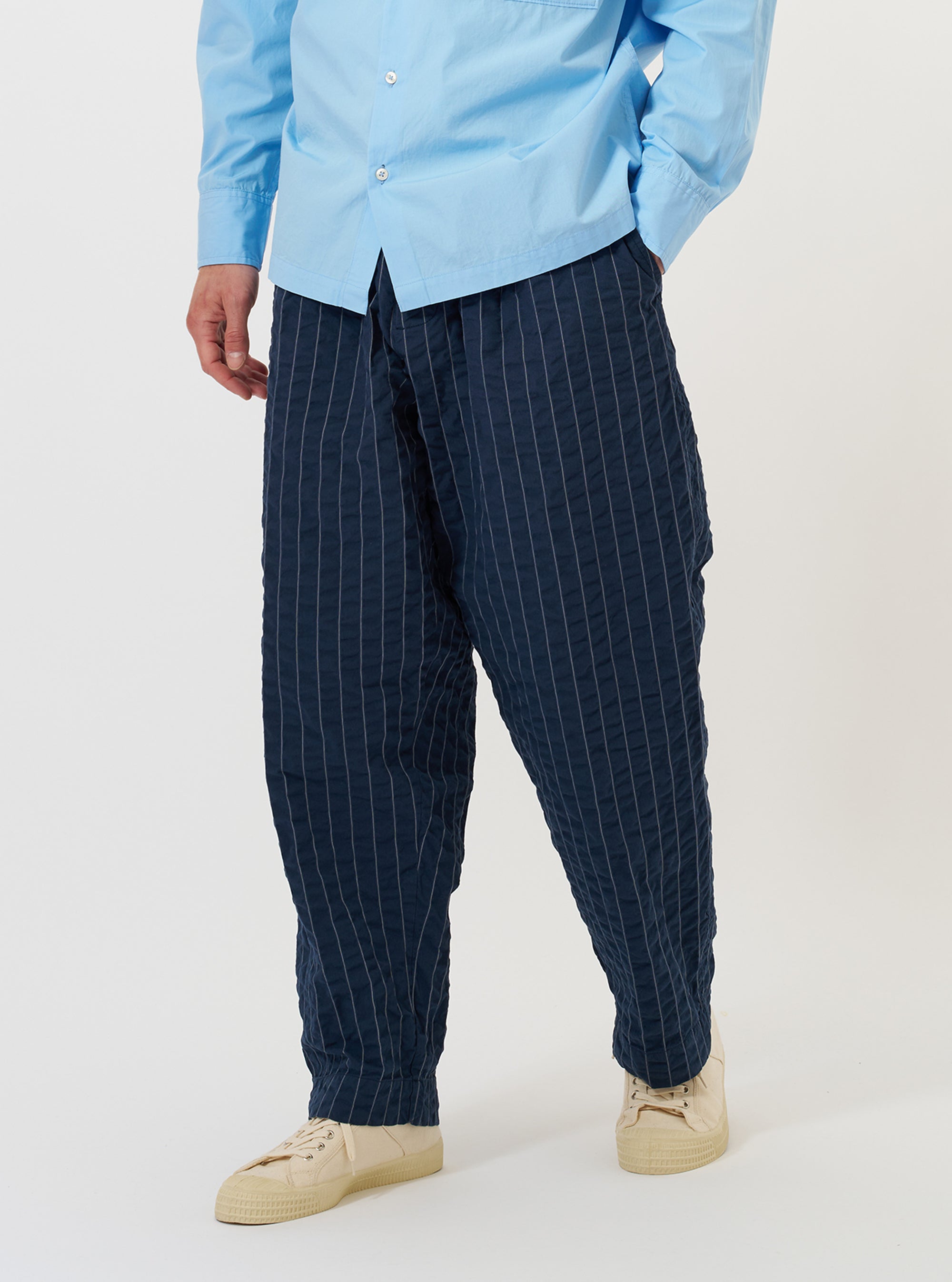 A model is wearing the Navy Chester Stripe Oxford Pant against a plain white background. The navy blue trousers feature a subtle vertical stripe pattern, a loose, wide-leg fit with a pleated front, and a drawstring visible at the waistband. The image shows the model from the waist down.