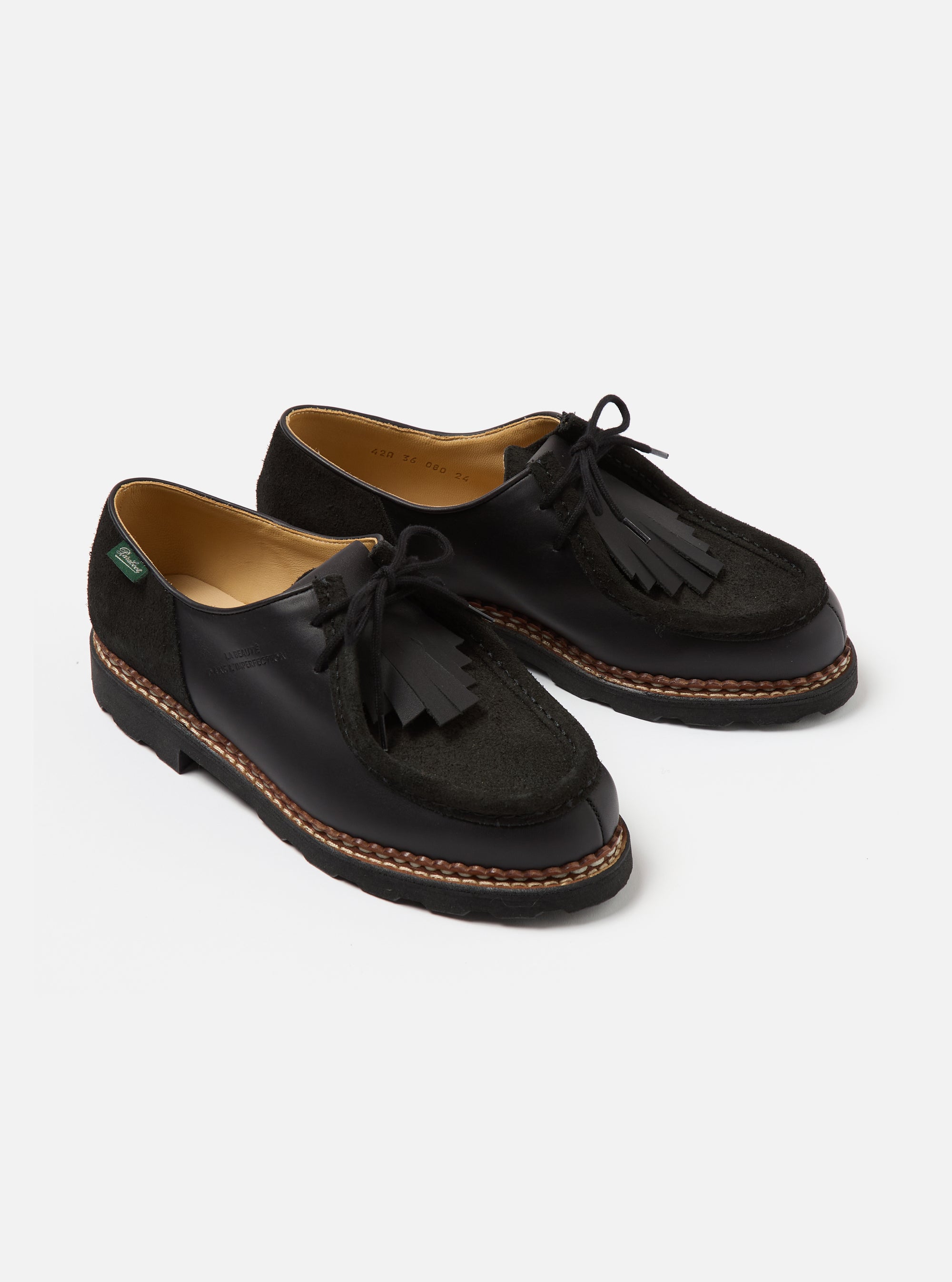 Paraboot x Universal Works Michael shoes in black leather and suede on a white background. The shoes have a fringe detail and a thick sole.