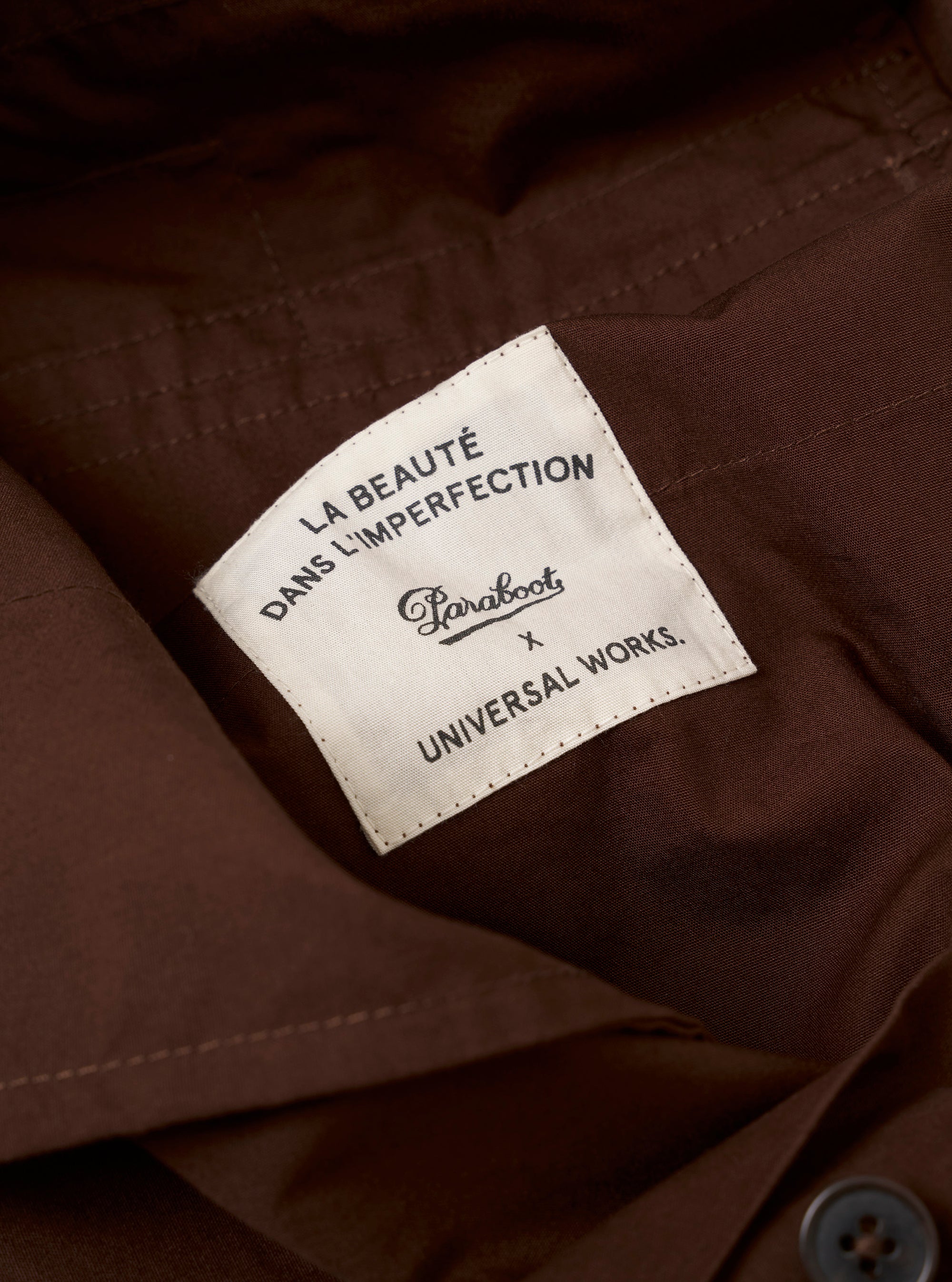 The Paraboot x Universal Works Brown Peached Poplin Super Chino is shown from a rear angle as a flat lay against a plain white background. The image highlights the two rear welt pockets, a small collaborative brand label above the right pocket, and the smooth, peached texture of the brown cotton poplin fabric.