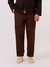 A model wears the Paraboot x Universal Works Brown Peached Poplin Super Chino against a plain, light grey background. The image is a full-length shot showing the model standing and facing forward, highlighting the trousers' relaxed, wide-leg fit and rich brown color. The chinos feature a single pleat at the front and a slightly cropped length with a turn-up cuffed hem. The model is wearing the trousers with a pair of dark shoes and a light-colored top.