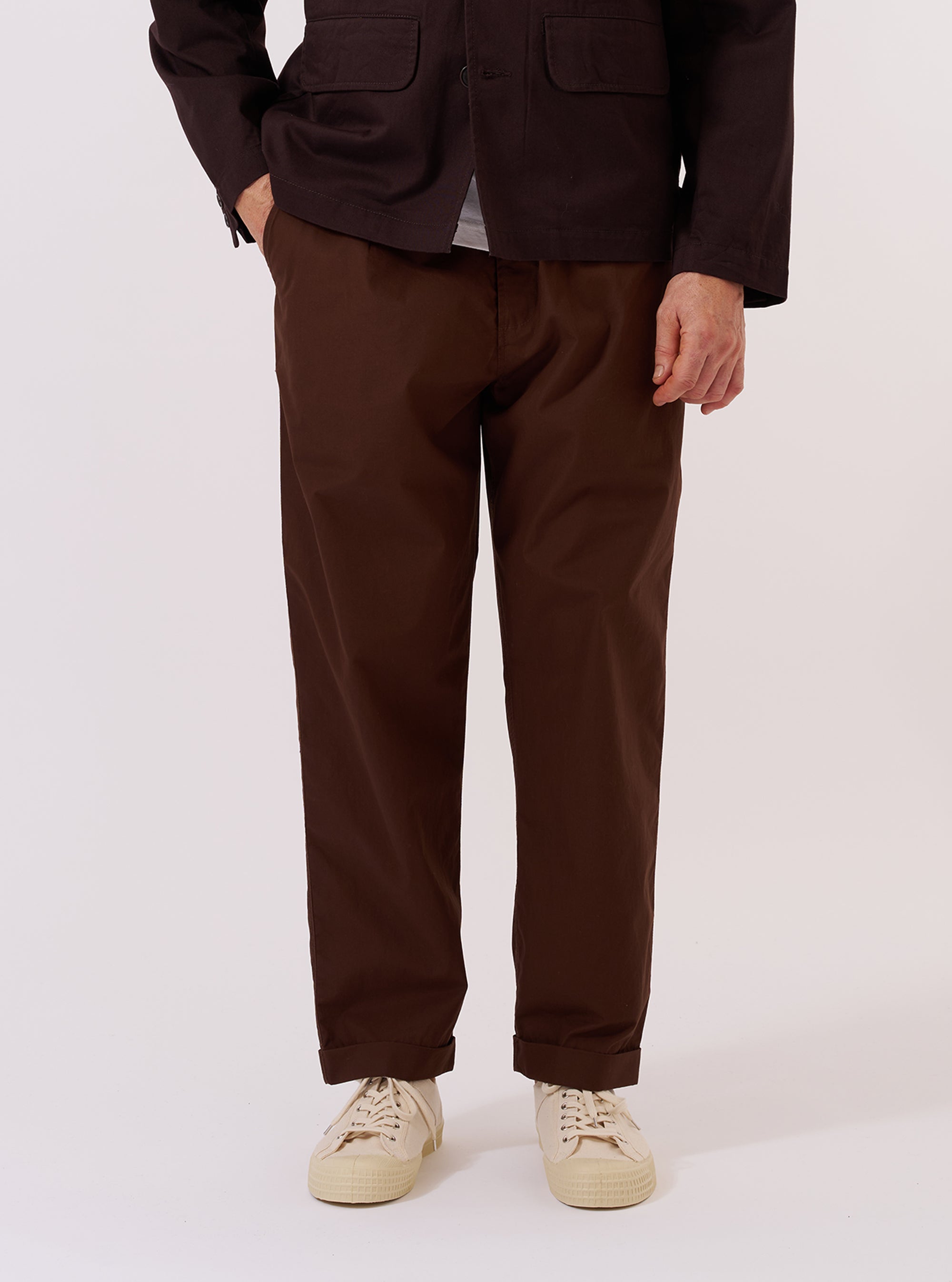 A model wears the Paraboot x Universal Works Brown Peached Poplin Super Chino against a plain, light grey background. The image is a full-length shot showing the model standing and facing forward, highlighting the trousers' relaxed, wide-leg fit and rich brown color. The chinos feature a single pleat at the front and a slightly cropped length with a turn-up cuffed hem. The model is wearing the trousers with a pair of dark shoes and a light-colored top.