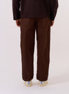 A model wears the Paraboot x Universal Works Brown Peached Poplin Super Chino, shown from a rear three-quarter angle against a plain white background. The image highlights the relaxed, straight-leg fit of the trousers and the smooth, matte texture of the brown peached poplin fabric. The model is wearing the chinos with dark shoes and a white T-shirt.