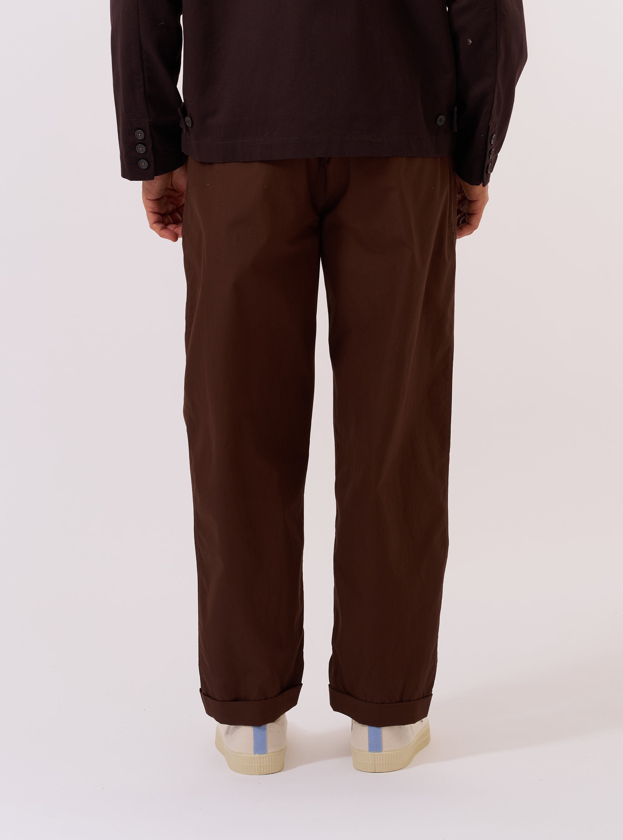 A model wears the Paraboot x Universal Works Brown Peached Poplin Super Chino, shown from a rear three-quarter angle against a plain white background. The image highlights the relaxed, straight-leg fit of the trousers and the smooth, matte texture of the brown peached poplin fabric. The model is wearing the chinos with dark shoes and a white T-shirt.