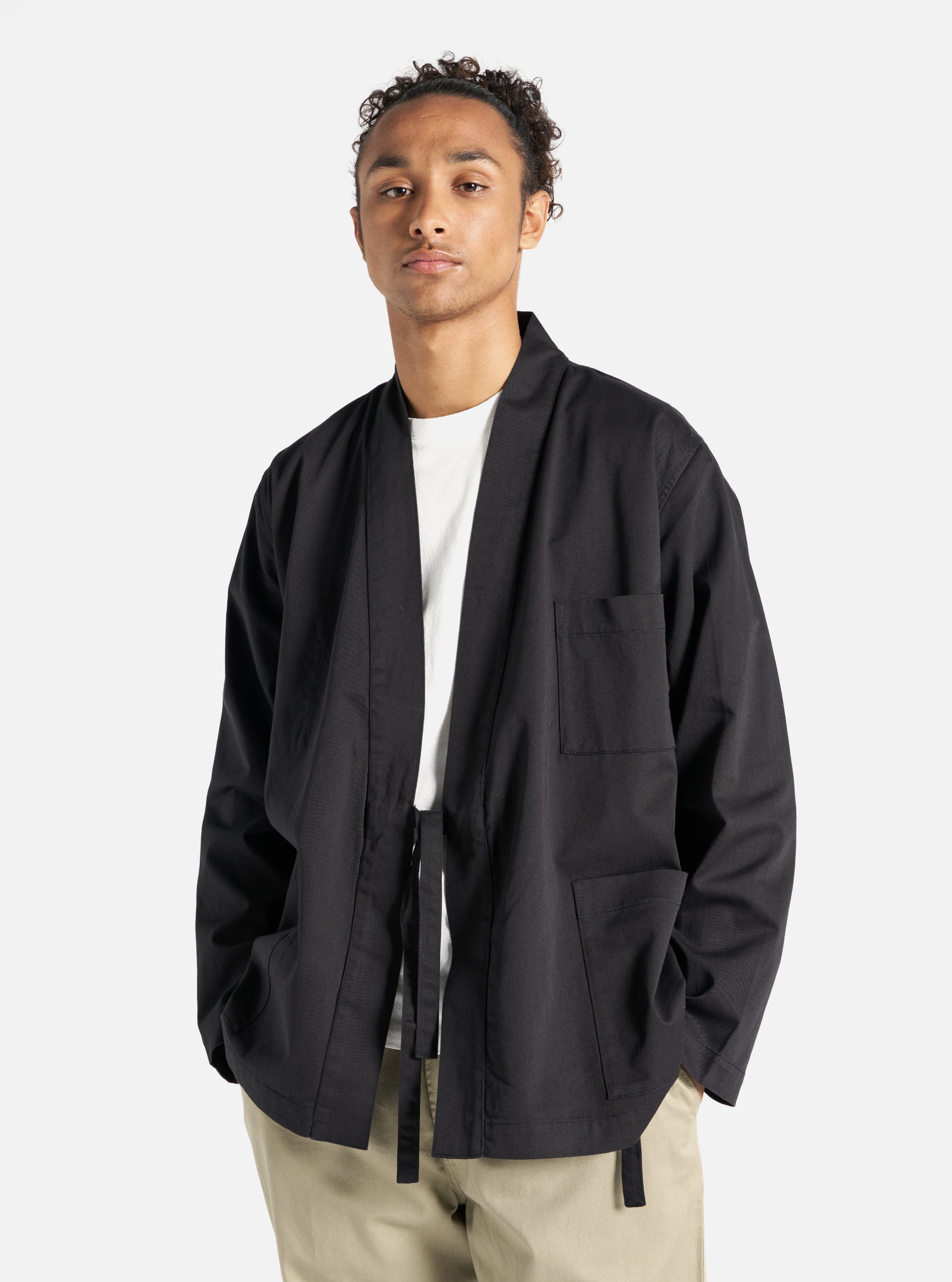 A model poses in a Black Twill Kyoto Work Jacket against a white backdrop. The jacket is black twill with a tie closure. He wears a white t-shirt and khaki pants.