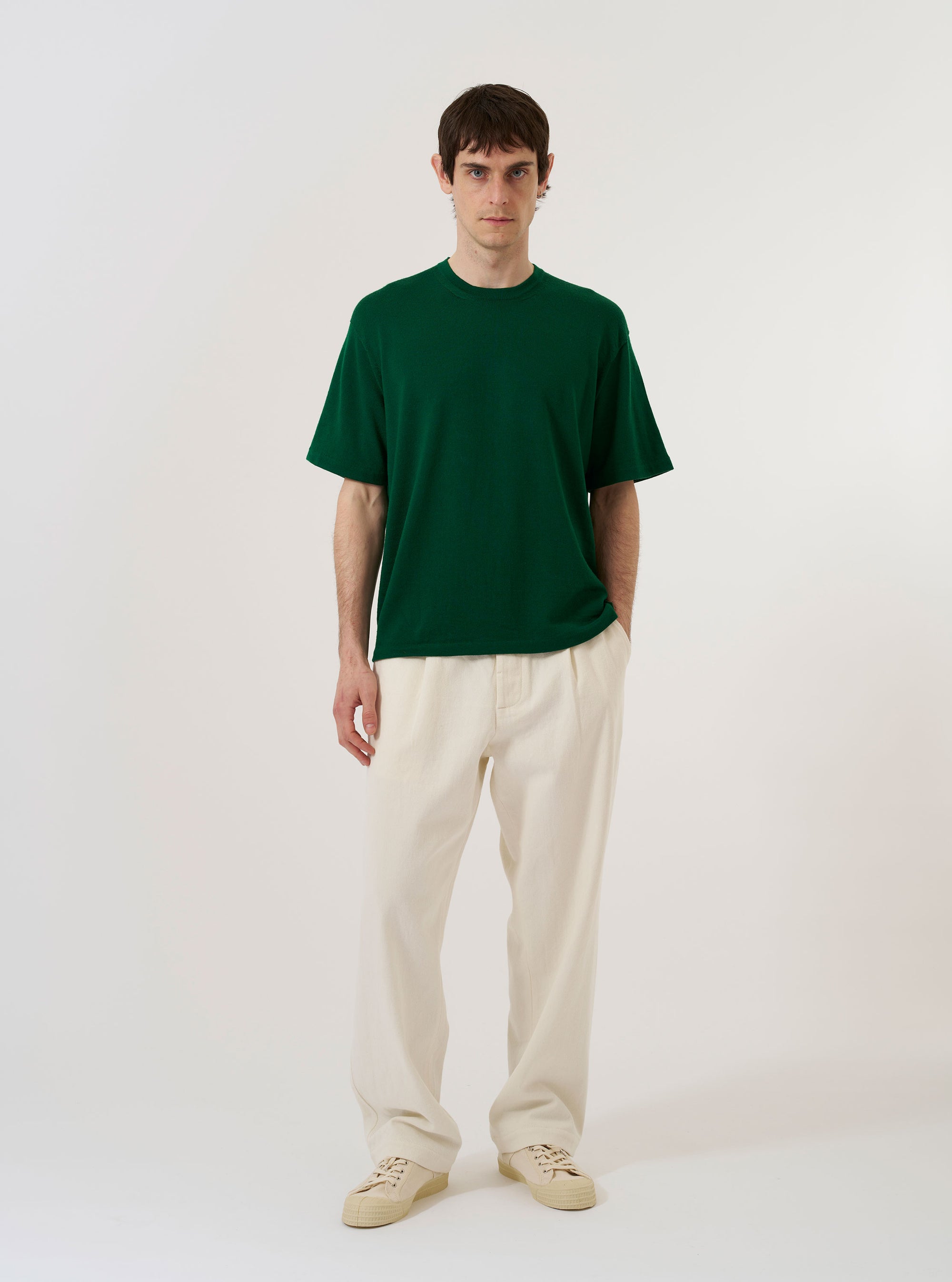 A model wears the Green Merino Fine Knit Knit Tee against a white background. The model is seen from the chest up, highlighting the fine knit texture, ribbed crew neck, and short sleeves of the green merino wool garment. The tee has a clean, minimalist fit.