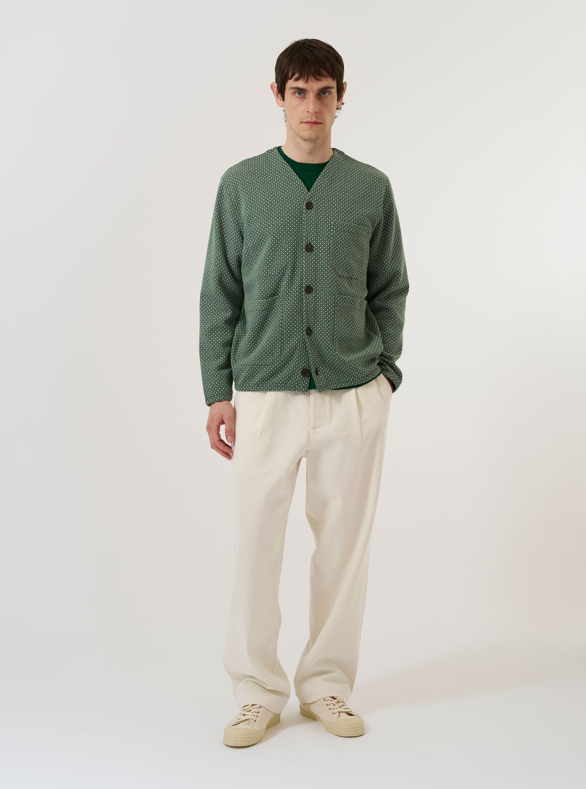 The Green Argyll Jersey Cardigan is worn by a model against a plain white background. The cardigan features a classic V-neckline, a central button-down closure with tonal buttons, and an all-over argyle pattern in shades of green. The model is wearing the cardigan layered over a light-colored collared shirt, highlighting the garment's relaxed fit and ribbed cuffs and hem.