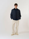 A model wears the Navy Recycled Polytech Parachute Field Jacket over a light-colored shirt and olive trousers, standing against a plain white background. The jacket features a contrasting brown corduroy collar, a buttoned neck strap, and four large 3D patch pockets with button flaps on the front. The garment has a loose, utilitarian fit with a straight hem and adjustable button cuffs.